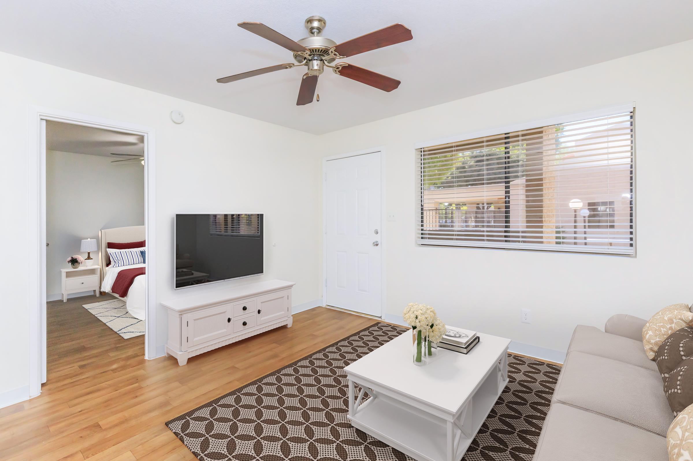 A bright and modern living room featuring a beige sofa, a coffee table, and a TV on a TV stand. There's a ceiling fan above and a window with blinds that lets in natural light. A doorway leads to another room, suggesting a cozy and inviting atmosphere. Wood flooring adds warmth to the space.