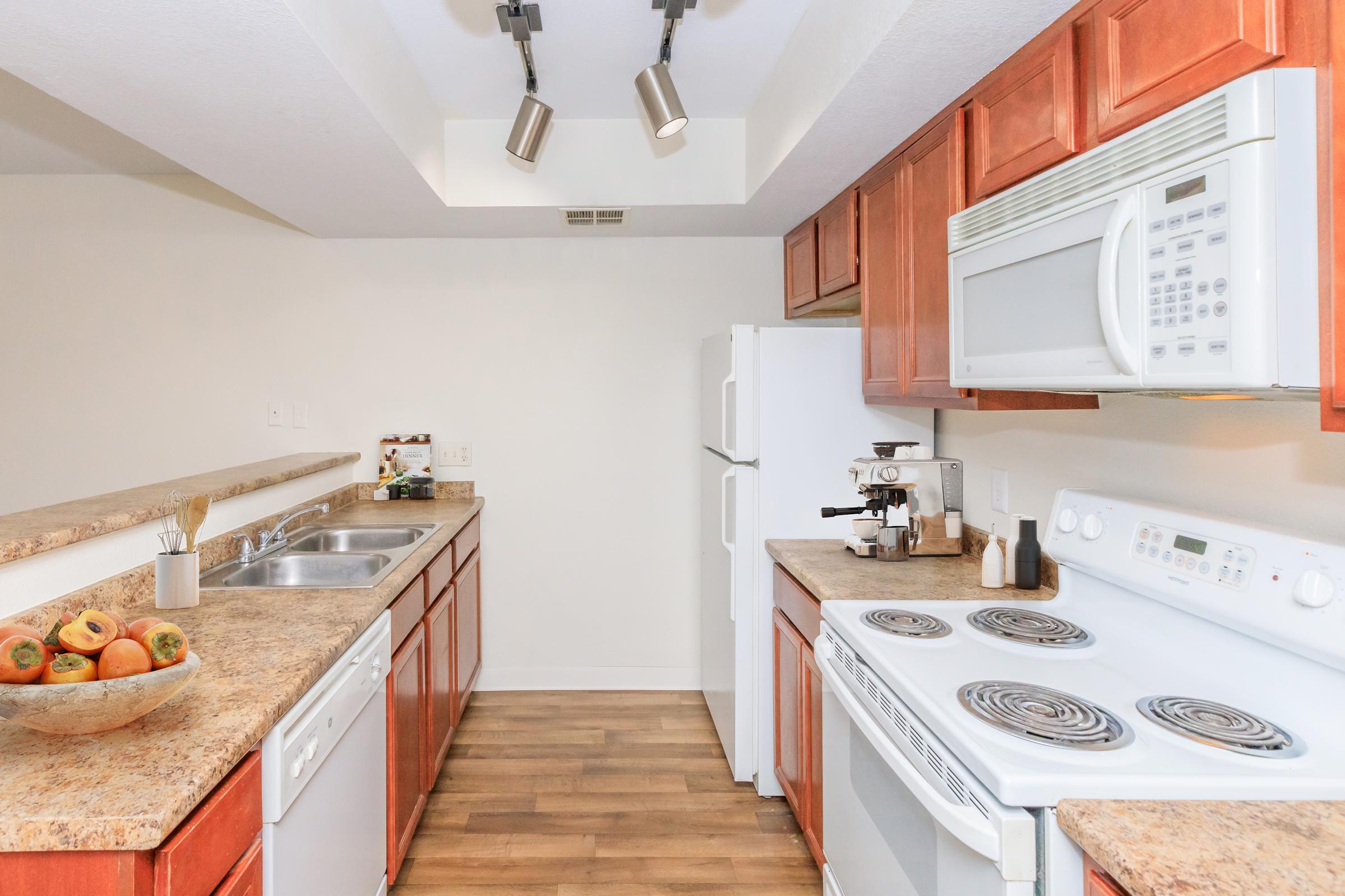A modern kitchen featuring wooden cabinets, a countertop with a sink, white appliances including a refrigerator and microwave, and a stove with an oven. The space is well-lit with track lighting, and a bowl of fruit is placed on the counter. The floor is covered with light wood laminate.
