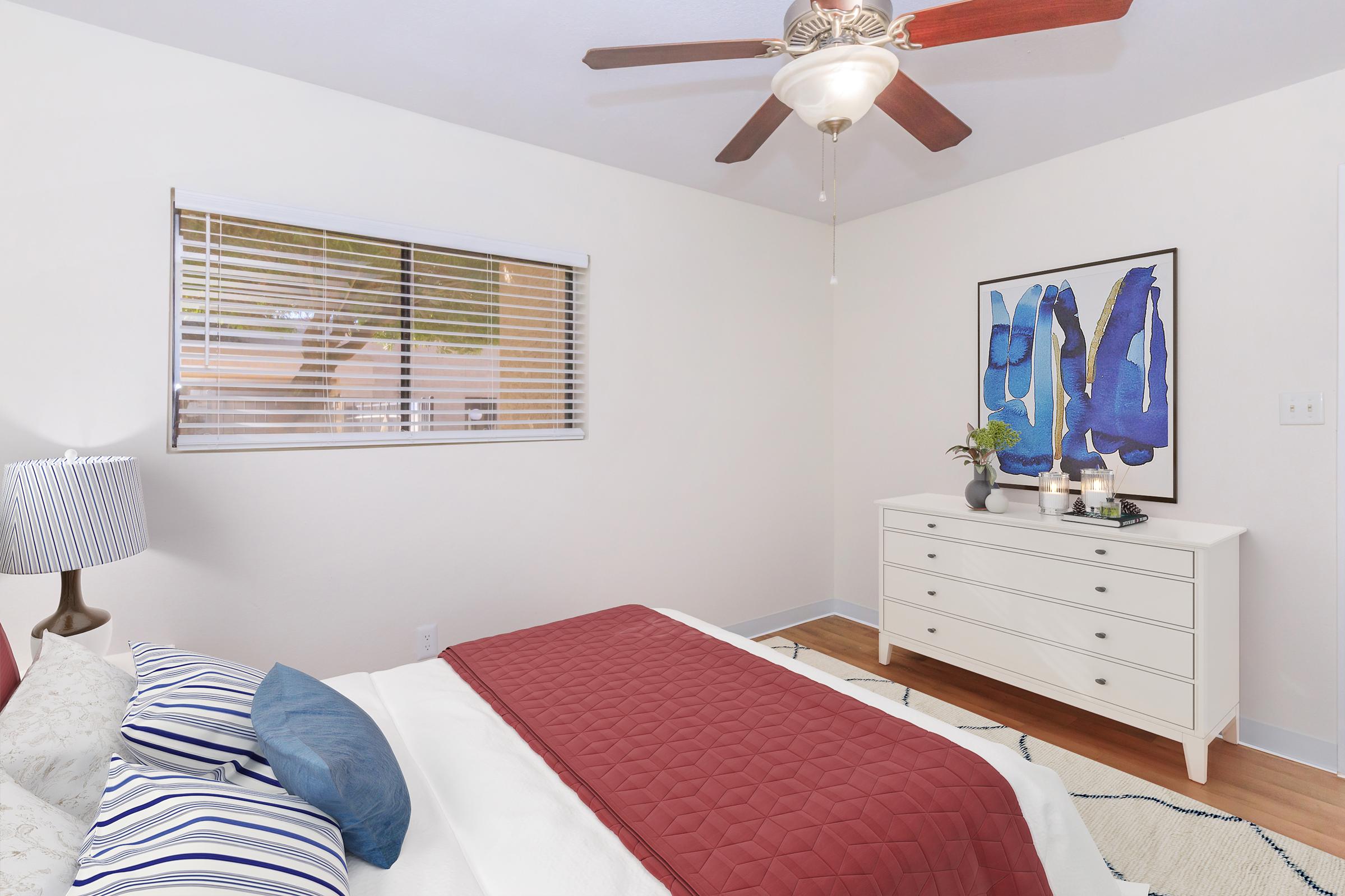 A cozy bedroom featuring a queen-size bed with a maroon quilt and decorative pillows. There is a white dresser against the wall, adorned with a small plant and decor. A large piece of abstract blue artwork hangs above the dresser, and a ceiling fan adds a modern touch. Natural light streams through a window with blinds.