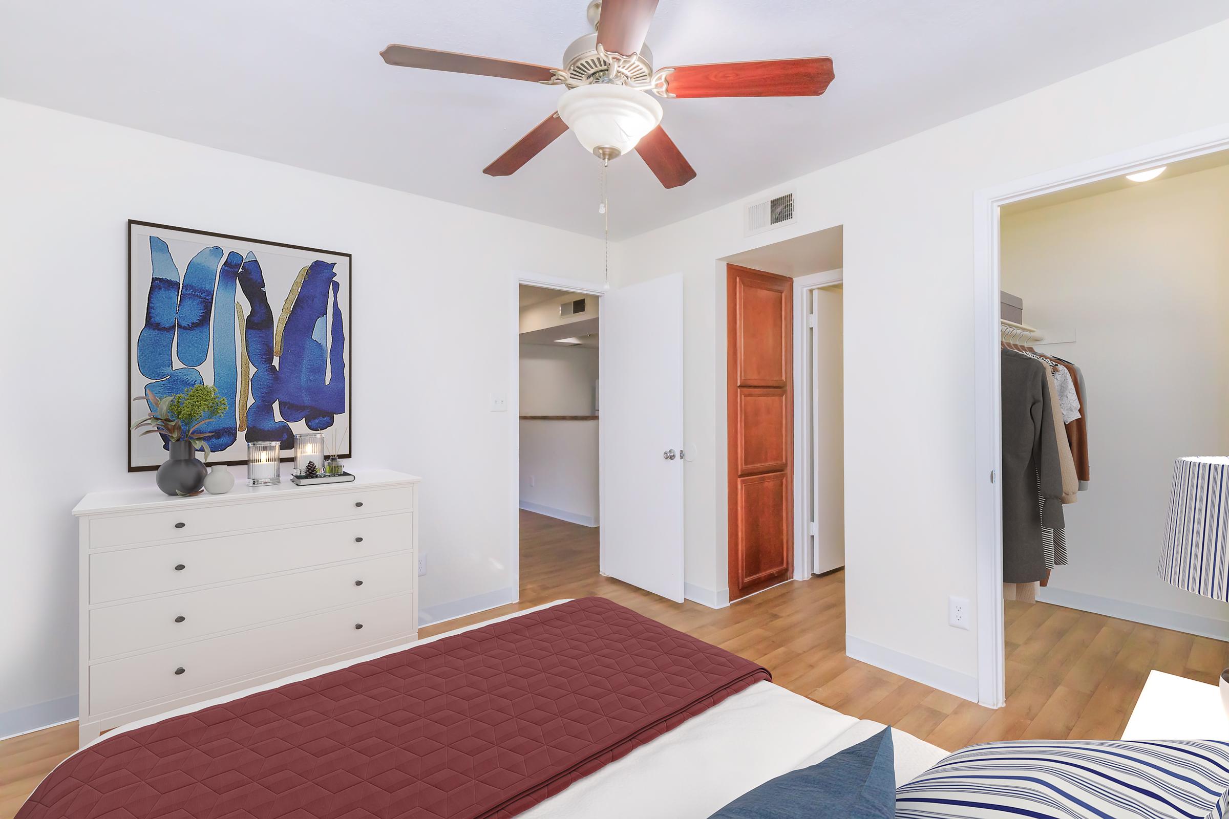 A bright and airy bedroom featuring a white dresser, a colorful framed artwork on the wall, and a ceiling fan with wooden blades. The bed has a burgundy coverlet and striped pillows. A closet with hanging clothes is visible in the background, enhancing the room's spacious feel.
