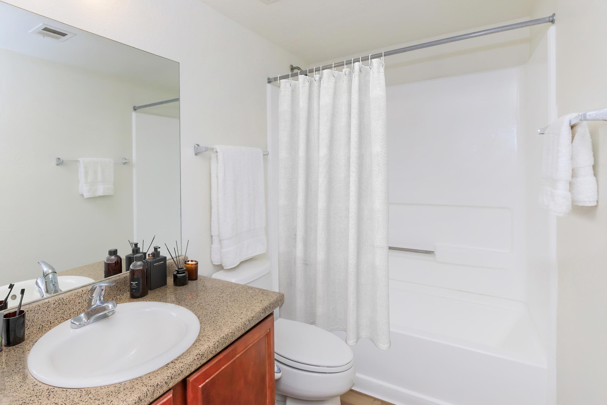 A clean and modern bathroom featuring a white bathtub with a shower curtain, a sink with a polished faucet, and a mirror above the sink. The countertop is adorned with various toiletries and two rolled towels. Light-colored walls and wooden flooring create a bright and inviting space.