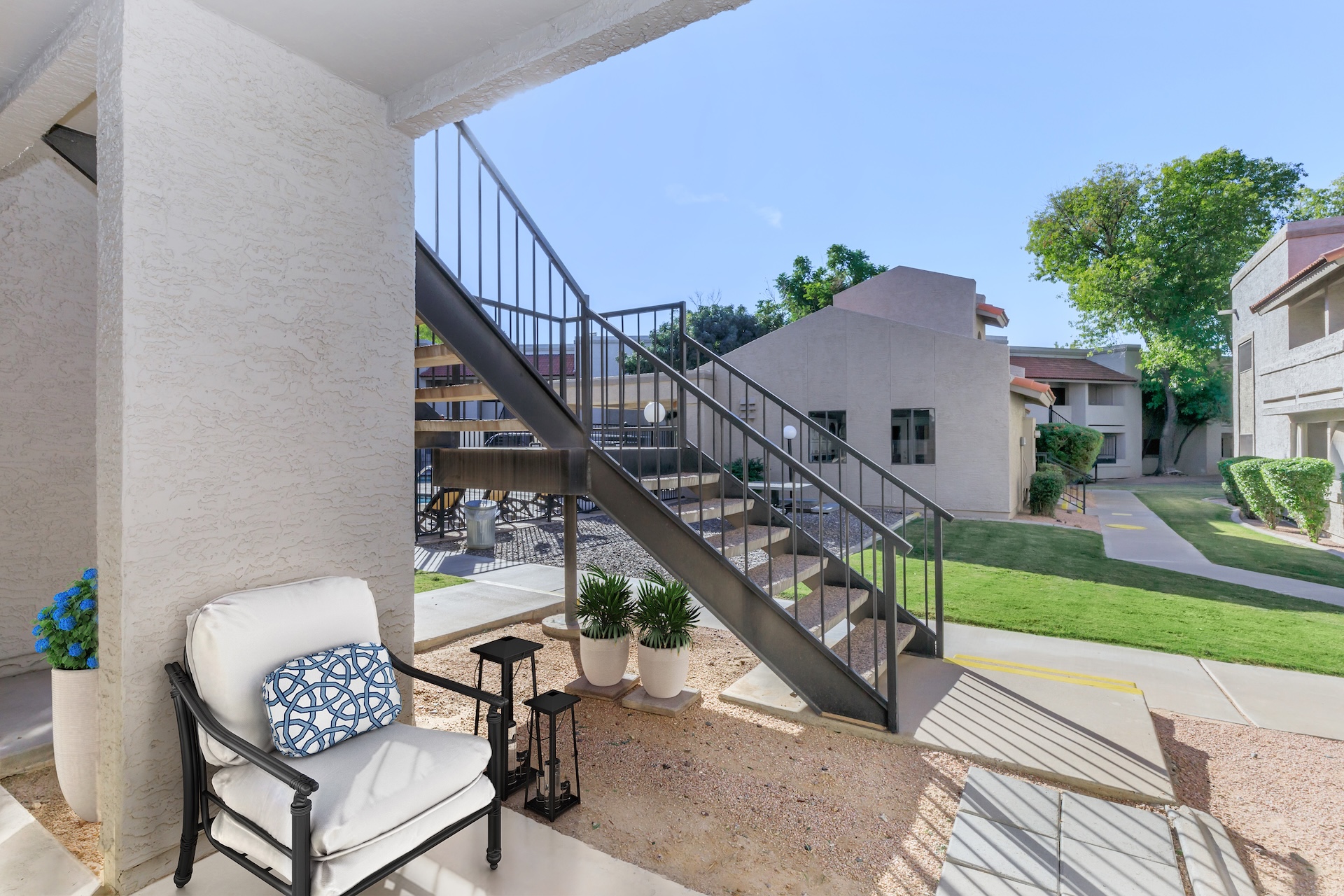 A view from a patio showing a metal staircase leading to the upper level of an apartment building. In the foreground, a chair with a decorative pillow and a small table are placed beside potted plants. Lush green lawns and additional apartment buildings are visible in the background under a clear blue sky.