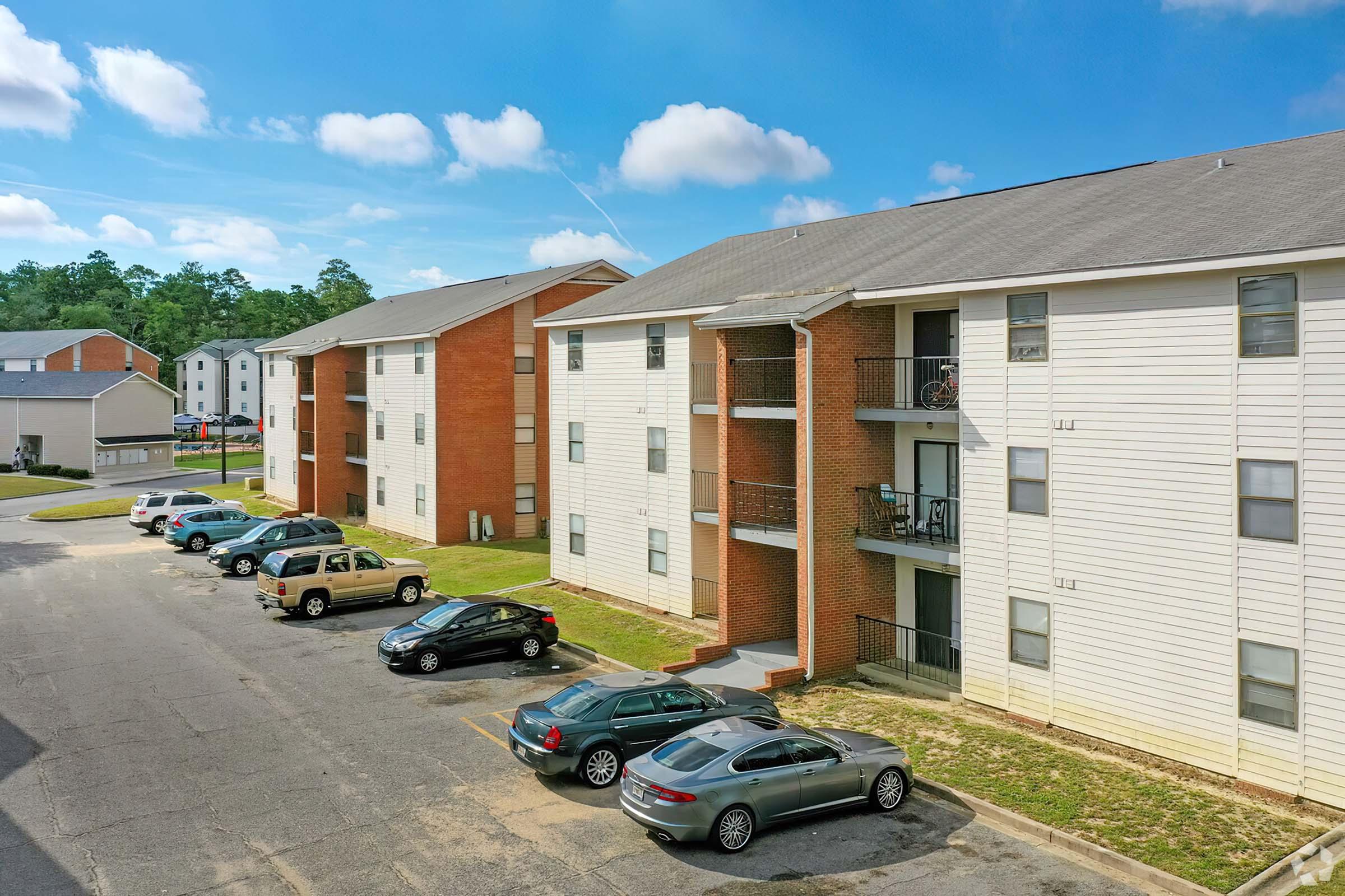 View of an apartment complex featuring three buildings with a mix of brick and siding exteriors. Several parked cars are visible in the foreground, and the scene is set under a blue sky with scattered clouds. The layout includes balconies on the buildings and grassy areas between the structures.