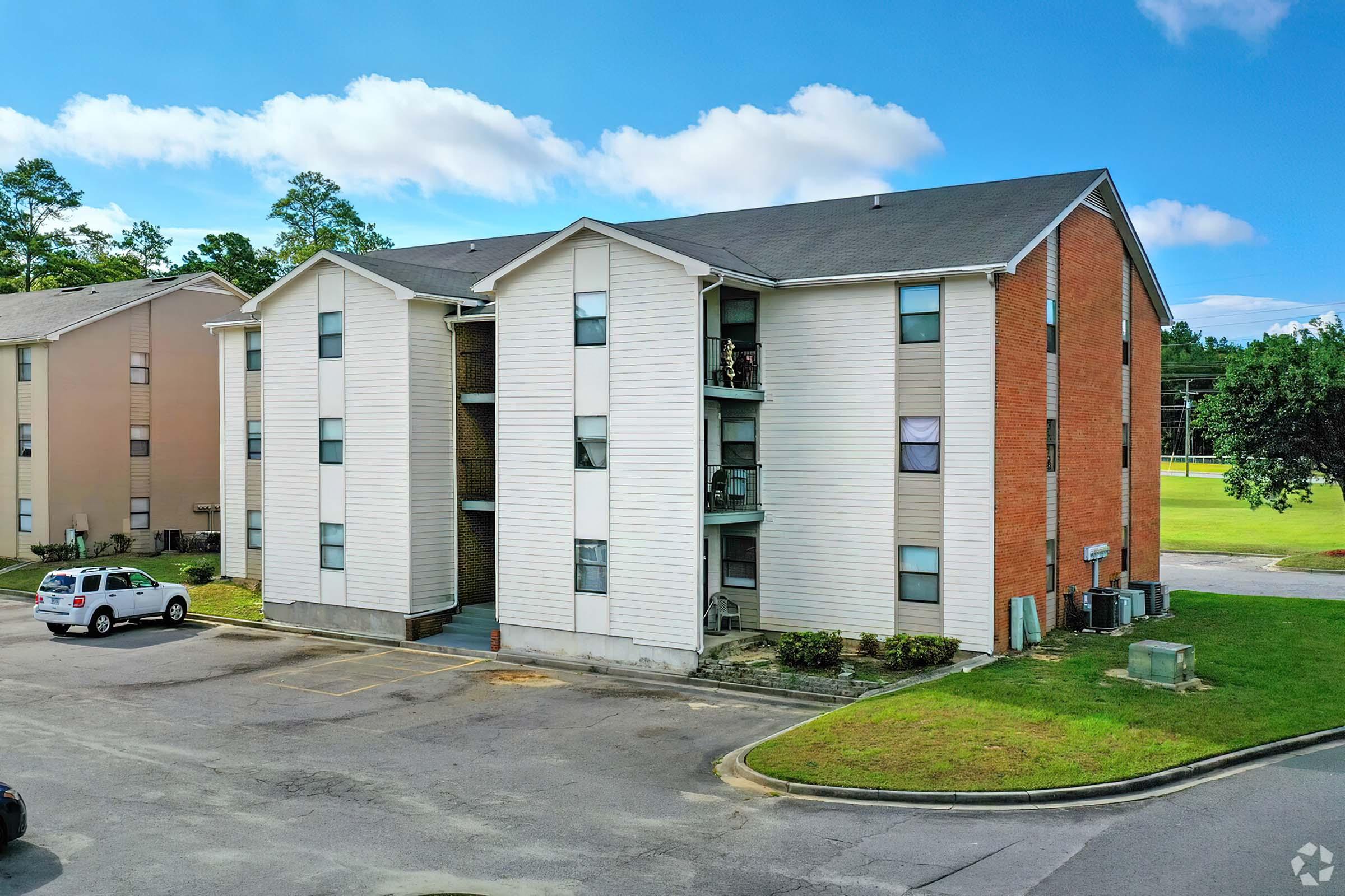 Exterior view of a two-story apartment building featuring a mix of white siding and brick. The building has multiple balconies and is surrounded by a paved parking area. Nearby greenery and trees are visible, along with an open space in the background.