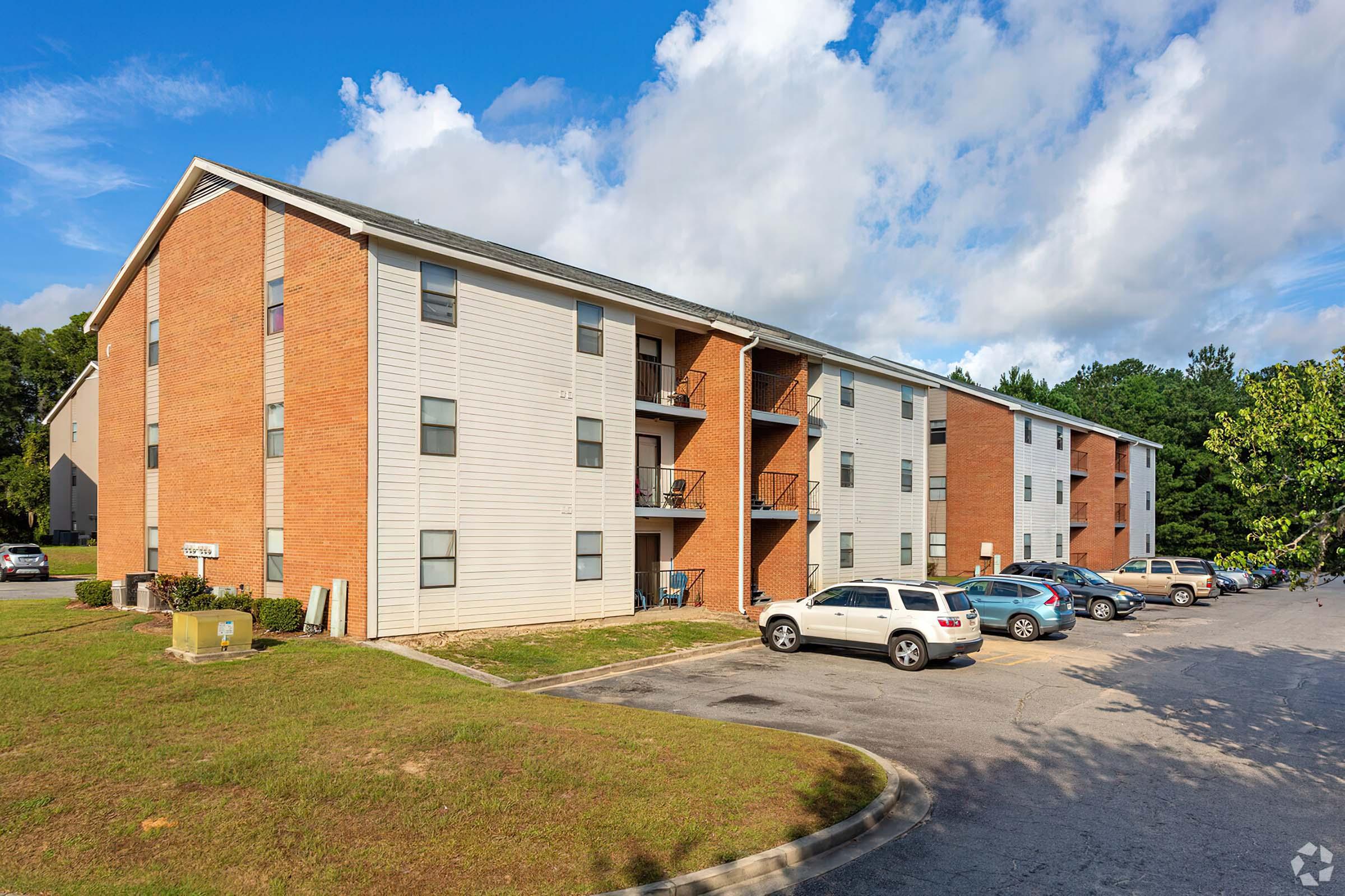 A multi-story apartment building with a combination of brick and siding exteriors. Several parked cars are visible in the foreground, and green trees line the background under a partly cloudy sky. The building features balconies on each level and has an open area in front.