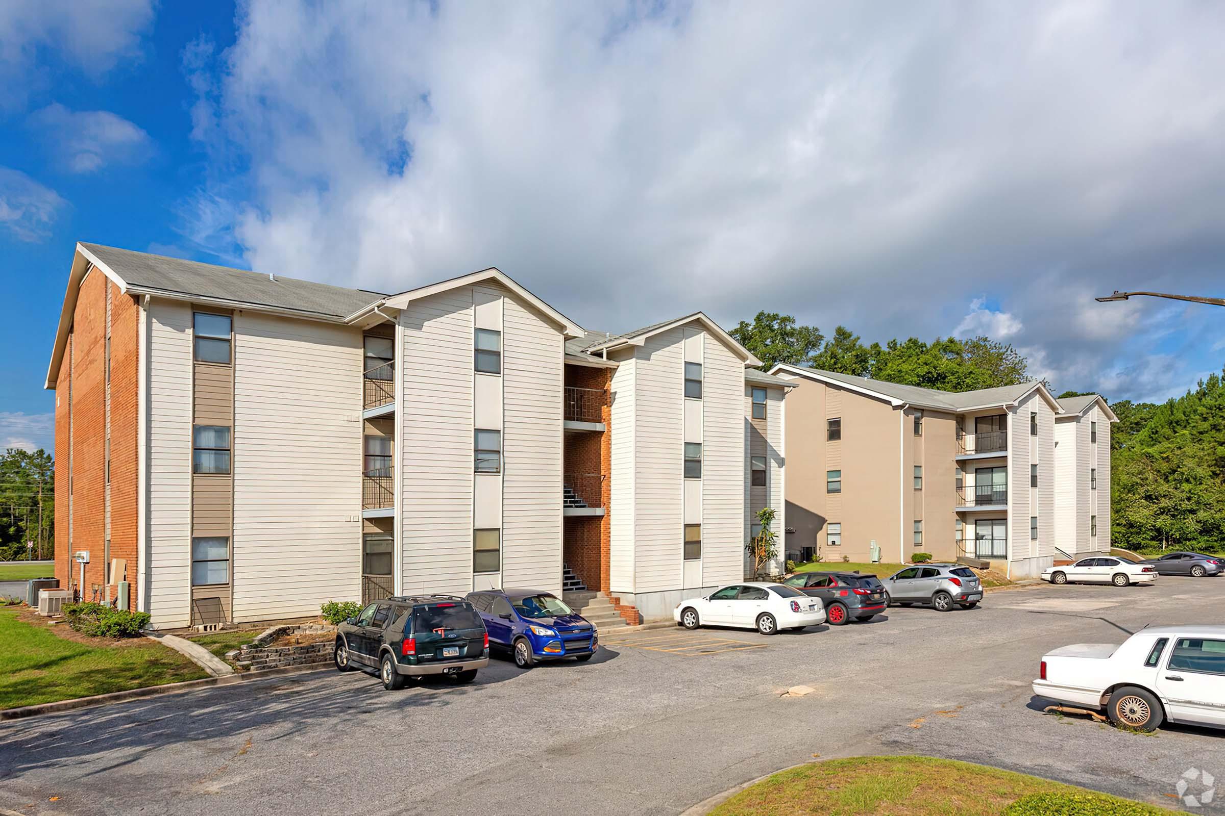 A three-story apartment complex featuring a mix of brick and siding exteriors, with several parked cars in the foreground. The scene is set against a backdrop of greenery and a partly cloudy sky.