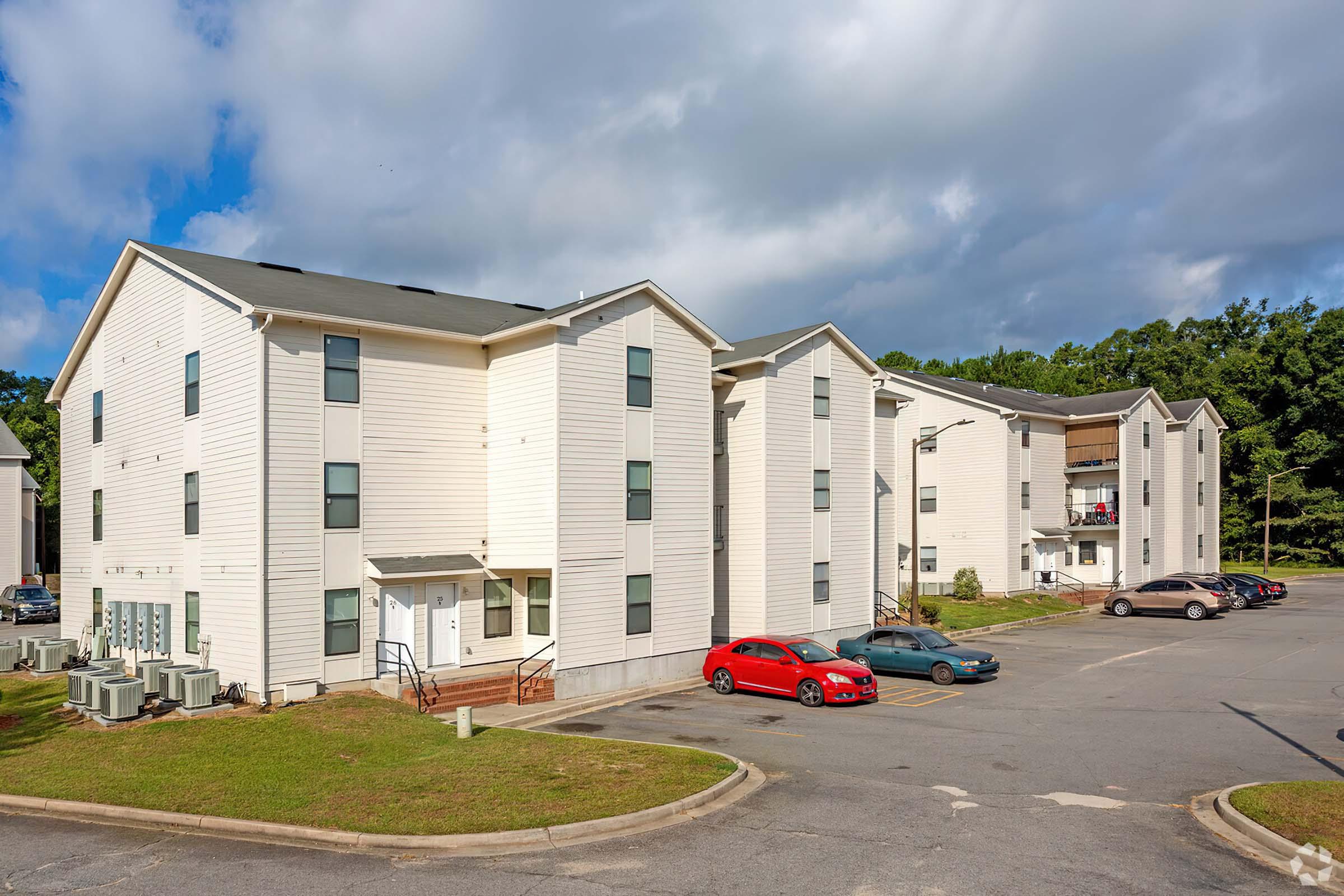 A view of a multi-unit residential building with three stories, featuring white siding and a parking lot. Several cars are parked in front, and air conditioning units are visible outside. The scene is set against a backdrop of a partly cloudy sky and green trees.