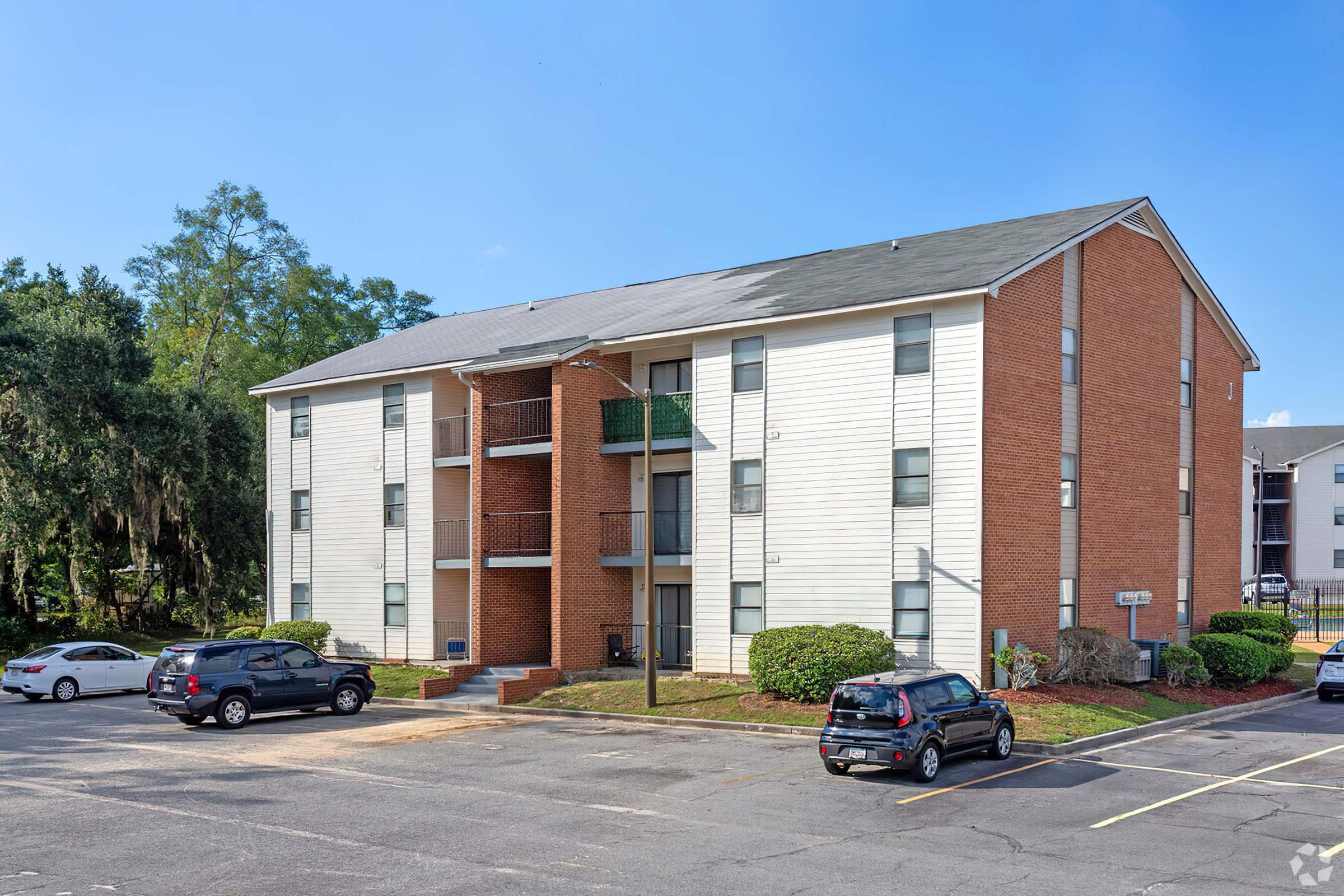 Three-story residential apartment building with a mix of brick and siding exterior. The building has balconies on the upper floors and accessible staircases. Surrounding it is a parking area with several parked cars and landscaped greenery. The sky is clear and blue.