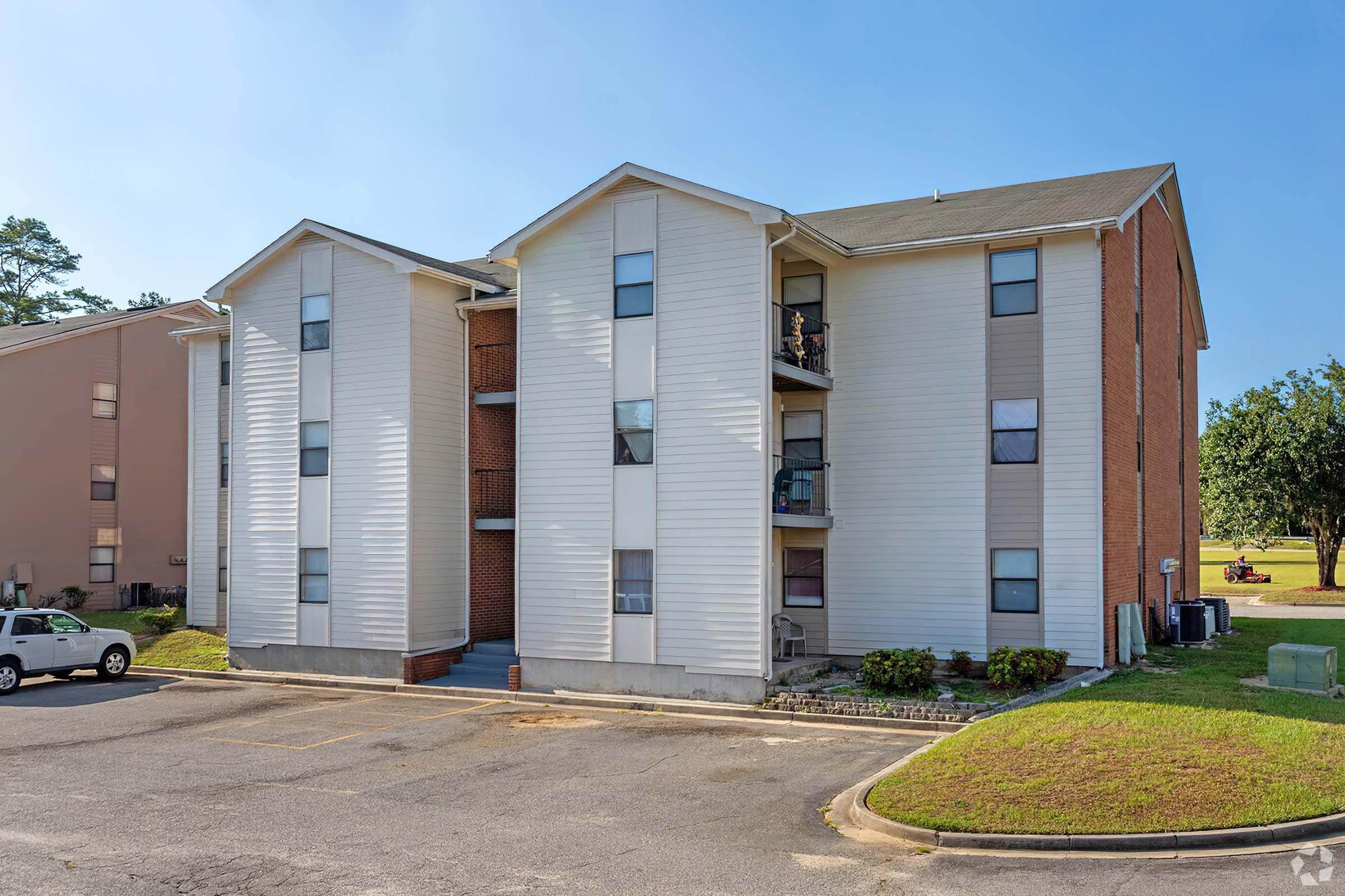 A three-story apartment building with a combination of brick and siding exterior. It features multiple balconies, some with items visible, and a small parking area in front. The surrounding area has grass and trees, indicating a residential setting. Clear blue skies are overhead.