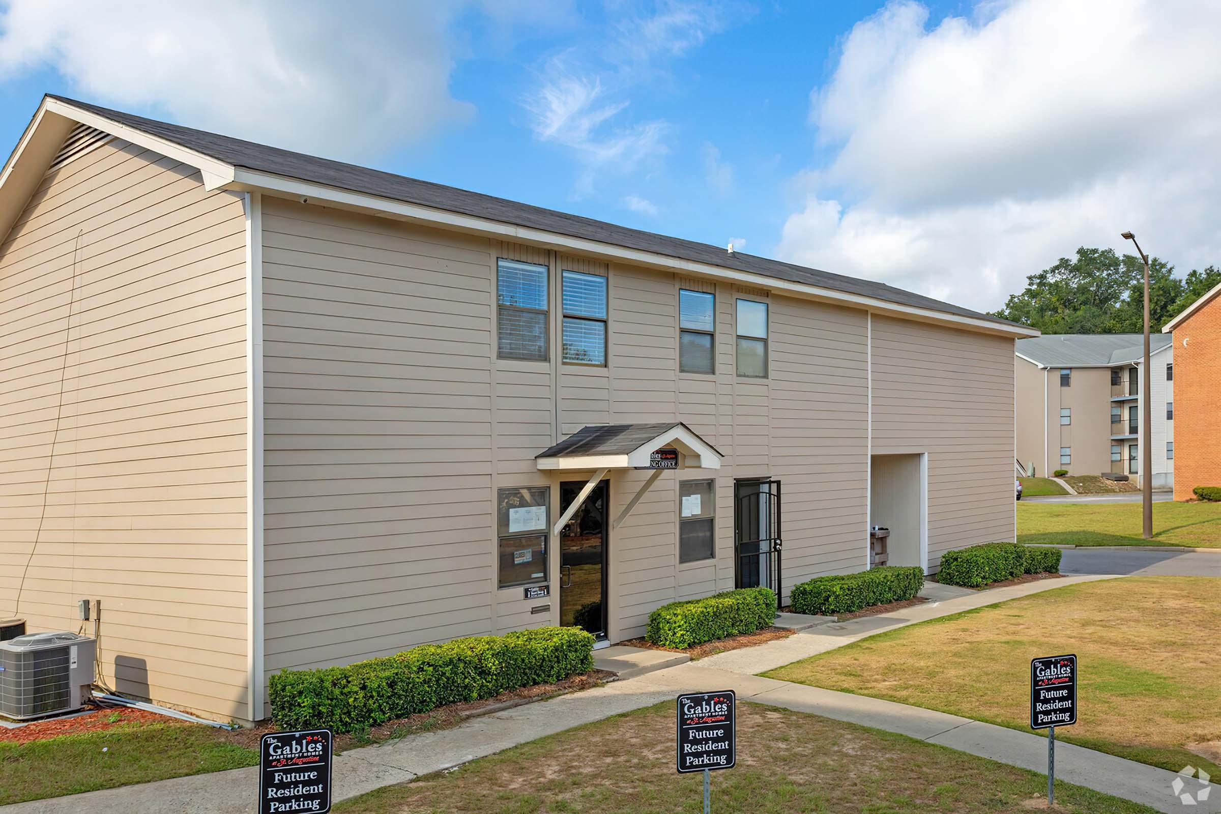 Two-story apartment building with a light-colored exterior and a black awning over the entrance. In front, there are neatly trimmed bushes and signs indicating reserved parking. The sky is partly cloudy, and there are additional buildings visible in the background.