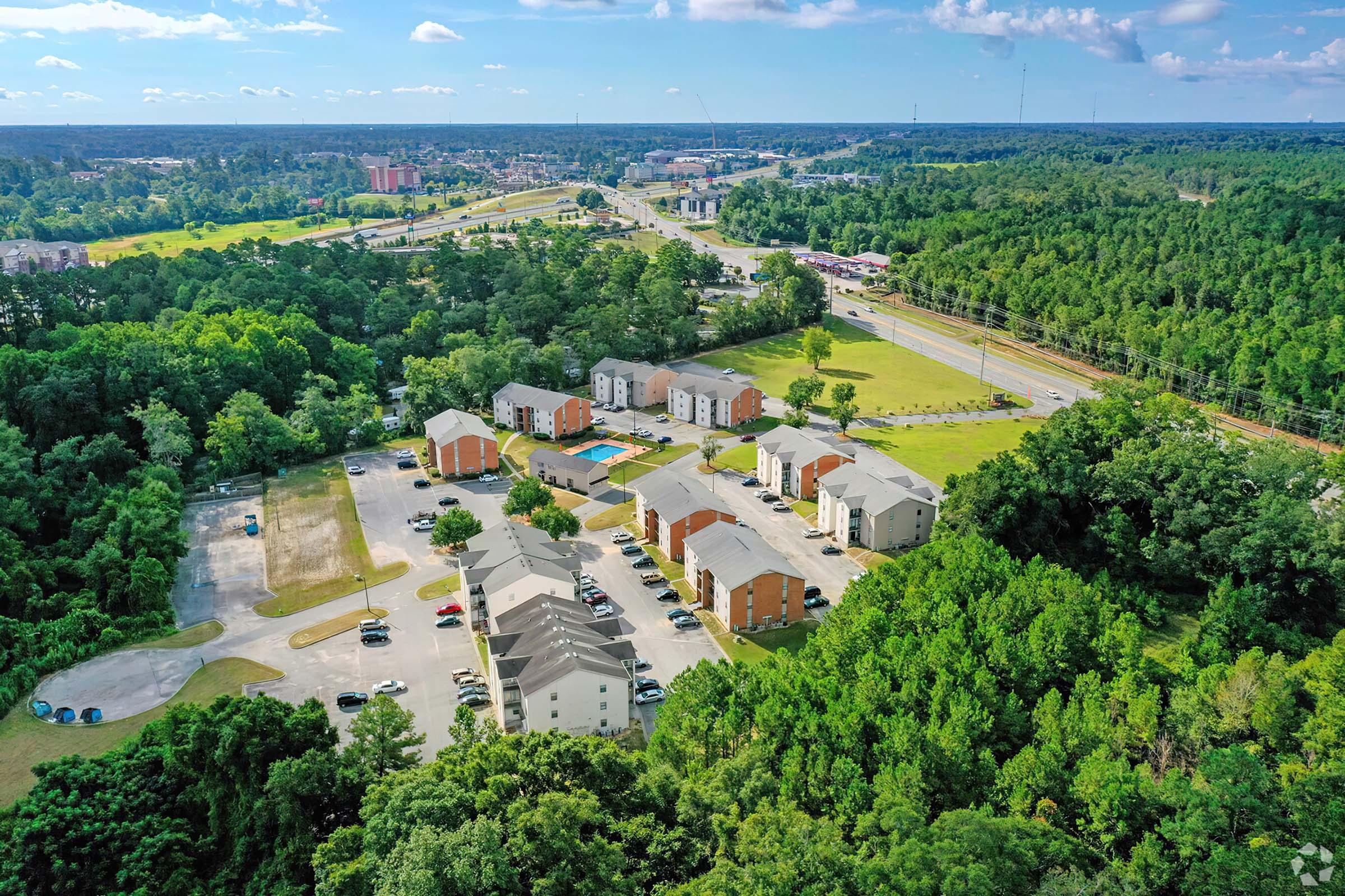 Aerial view of a residential complex surrounded by greenery, featuring several multi-story buildings and an outdoor pool. Nearby, a road and commercial area are visible in the background, along with patches of forested land. The sky is blue with scattered clouds.