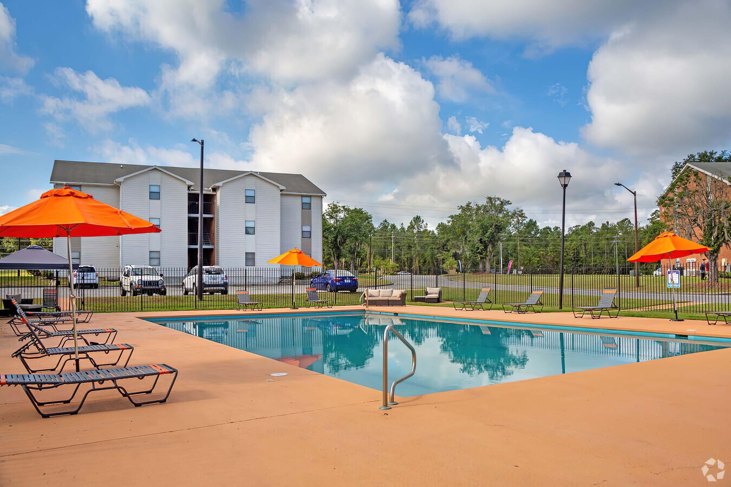 A sunny pool area featuring a clear blue swimming pool surrounded by lounge chairs and orange umbrellas. In the background, there are two-story apartment buildings and a grassy area, with a few cars parked nearby under a partly cloudy sky.