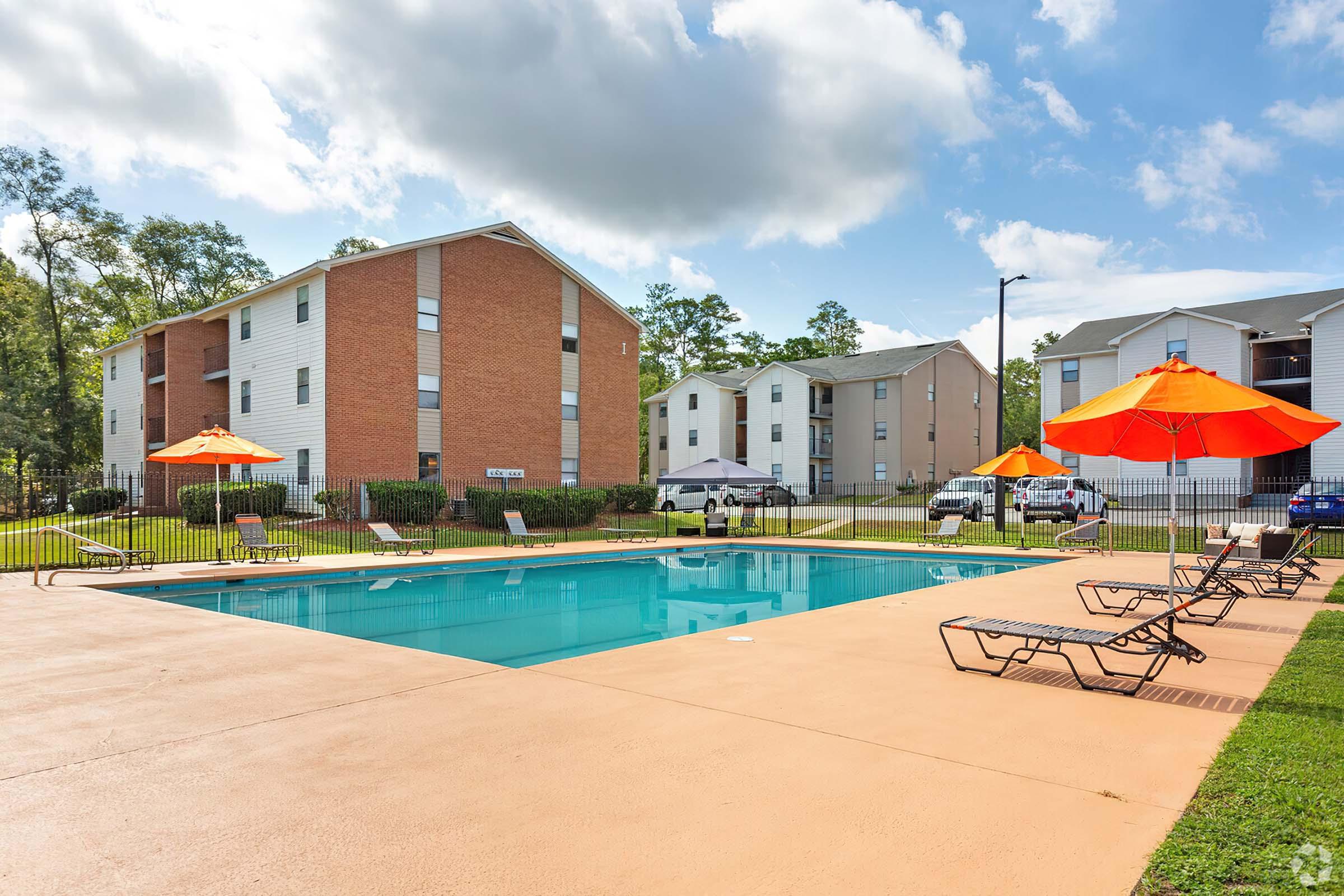 A serene outdoor pool area surrounded by lounge chairs and vibrant orange umbrellas. In the background, two multi-story residential buildings and a small parking area are visible against a clear blue sky with scattered clouds. The setting conveys a relaxed and inviting atmosphere.