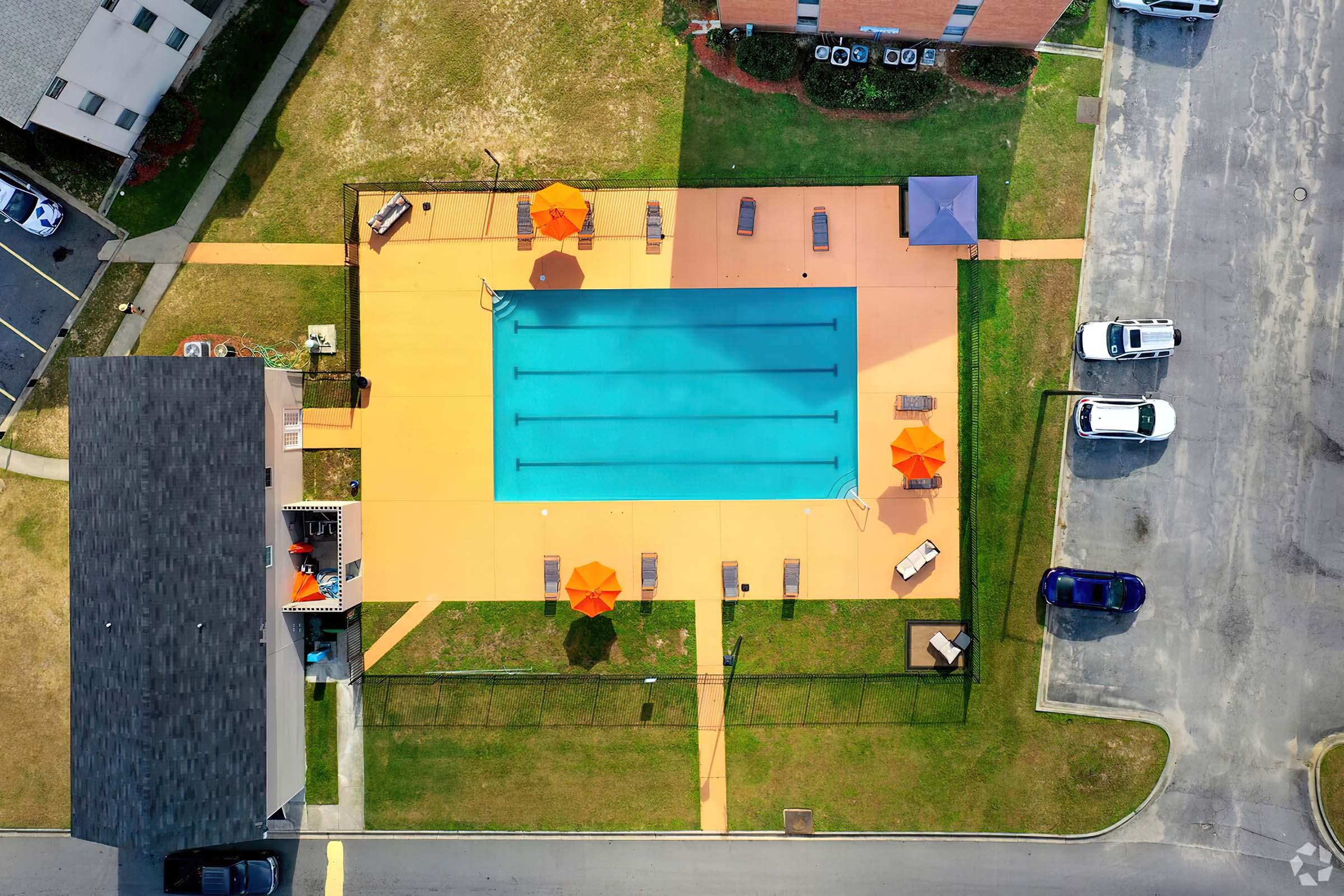 Aerial view of a swimming pool surrounded by orange umbrellas and lounge chairs. The pool features six lanes, with grassy areas and a paved surface surrounding it. Nearby, there are parked cars and two buildings, one with a dark roof and the other with a red facade.