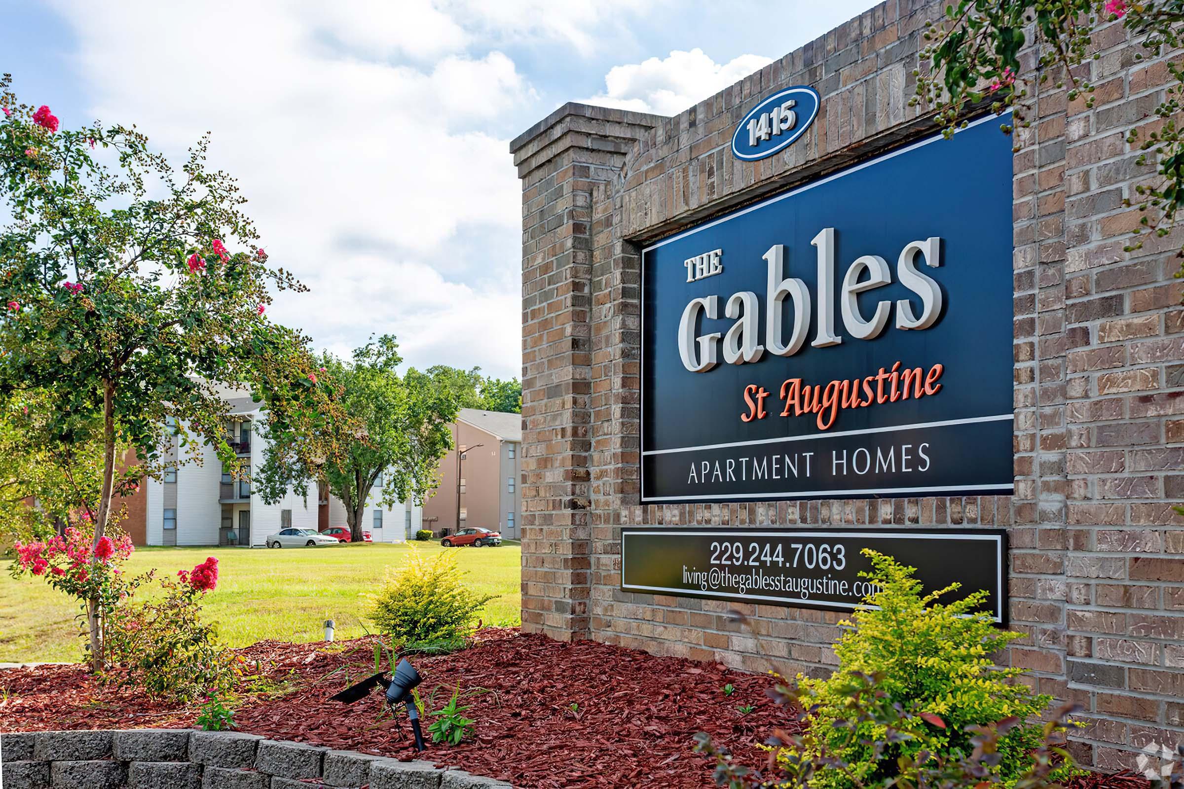 A sign for "The Gables St Augustine Apartment Homes," featuring the address "1415." Surrounding the sign are vibrant flowers and landscaping, with a clear blue sky in the background.