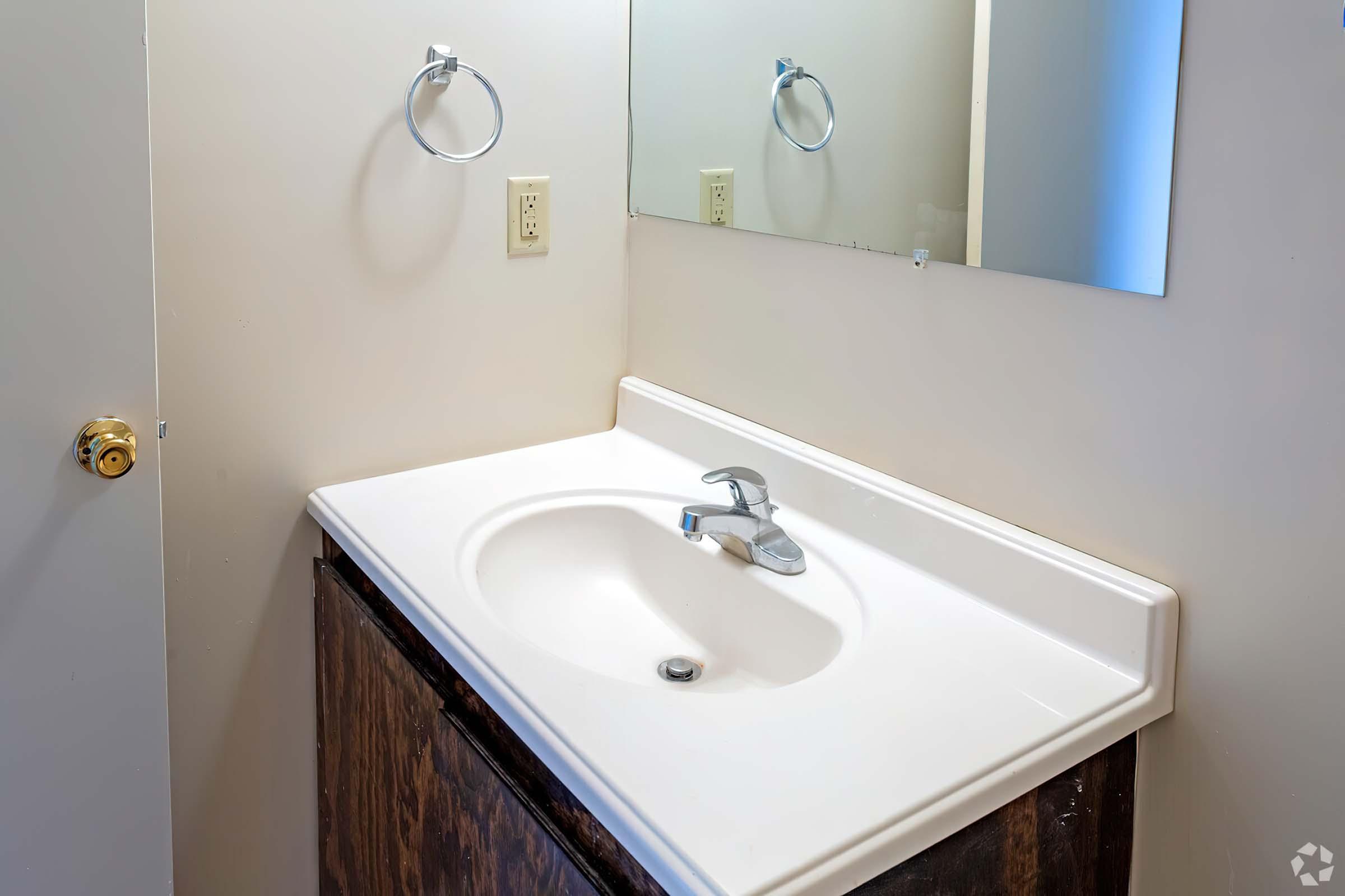 A bathroom sink with a single faucet sits on a countertop. The wall is bare, featuring two electrical outlets and a small mirror above the sink. The cabinet below is made of dark wood, and the overall lighting is bright and neutral.