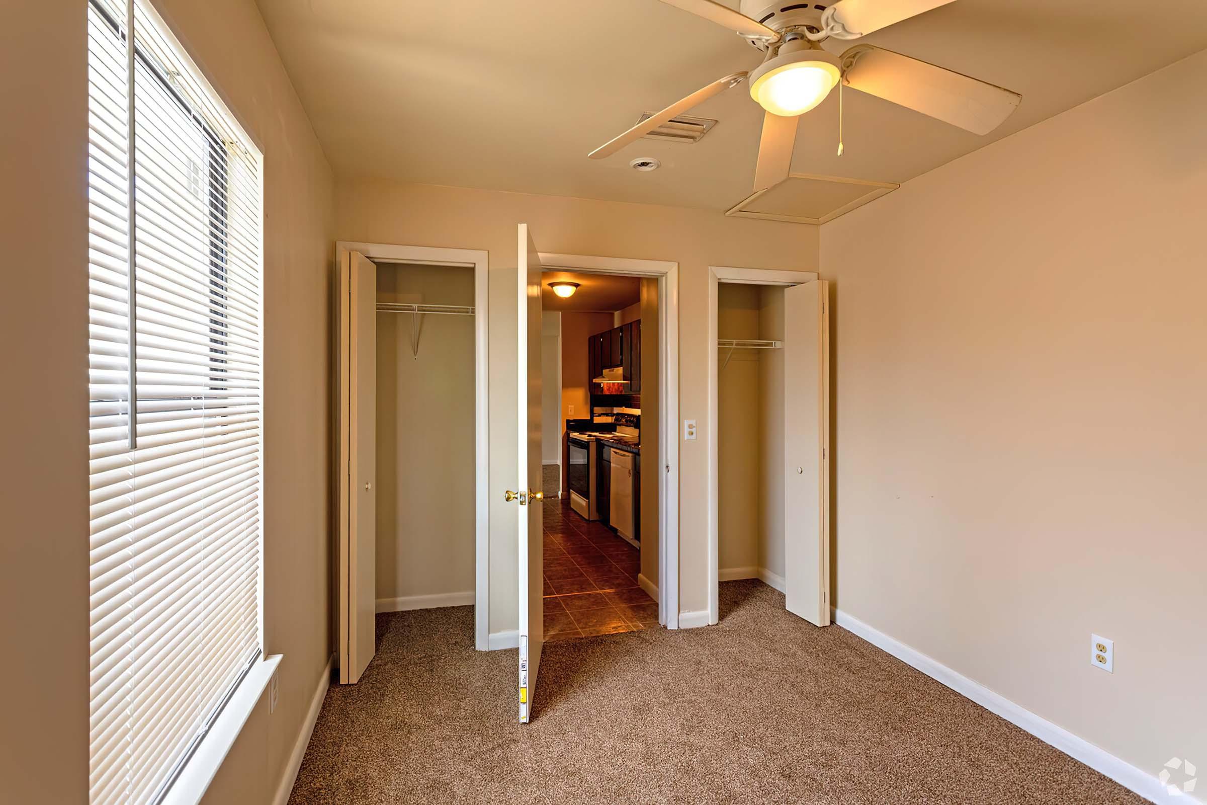 Interior view of a small room featuring light-colored walls, a ceiling fan, and carpeted flooring. Two open closet doors are visible on the left, while a doorway leads to a kitchen area at the back. A window with blinds allows natural light to enter, enhancing the warm ambiance of the space.