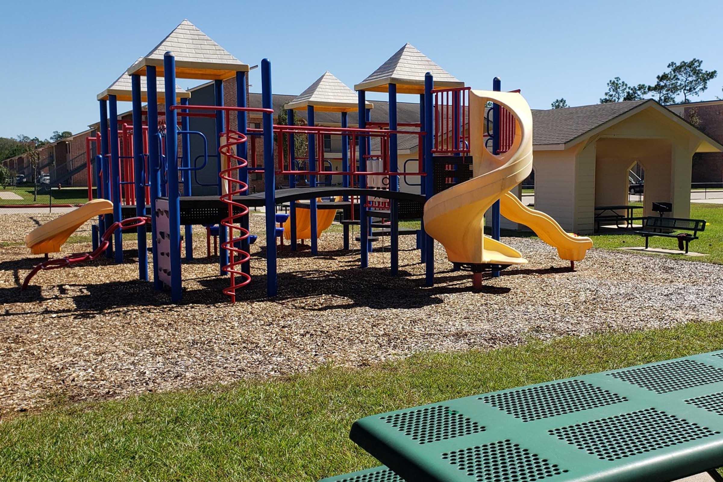Colorful playground equipment featuring slides, climbing structures, and monkey bars, situated on a bed of mulch. A bench is visible in the foreground with a clear blue sky and a building in the background. The scene suggests a sunny, inviting area for children to play.