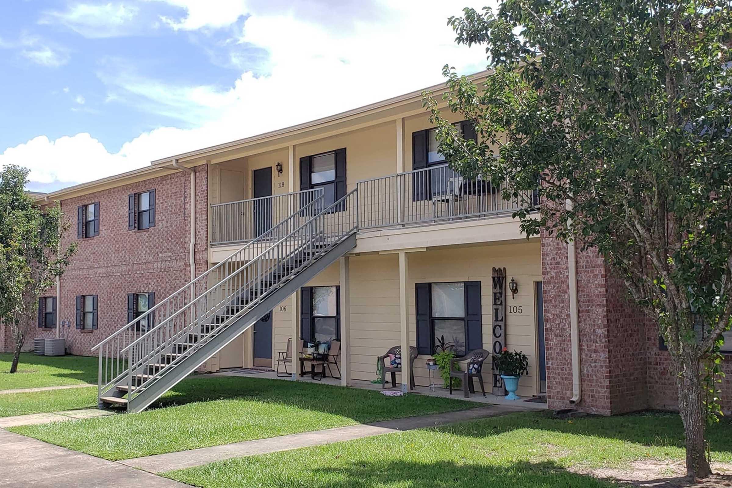 Two-story apartment building with a brick facade and beige accents. A set of gray stairs leads to an upper entrance, marked with a "Welcome" sign. Two chairs are placed on the porch, surrounded by green lawns and small trees on either side. The sky is partly cloudy, creating a bright atmosphere.