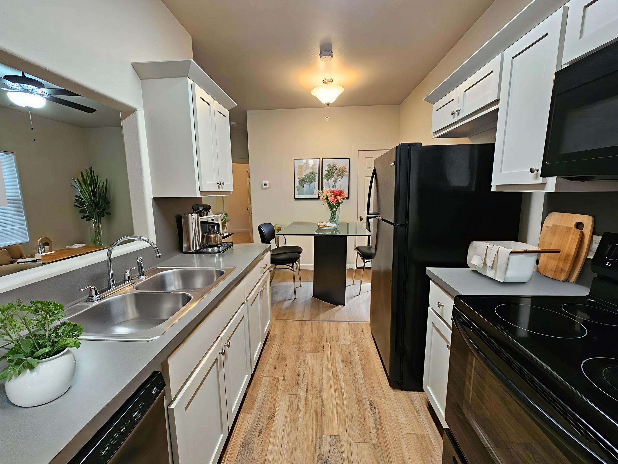 Bright, modern kitchen featuring white cabinets, a double sink, and stainless steel appliances. A black refrigerator and stove complement the light wood flooring. The adjacent dining area includes a glass table with chairs, and there's a small plant on the countertop, creating a welcoming atmosphere.