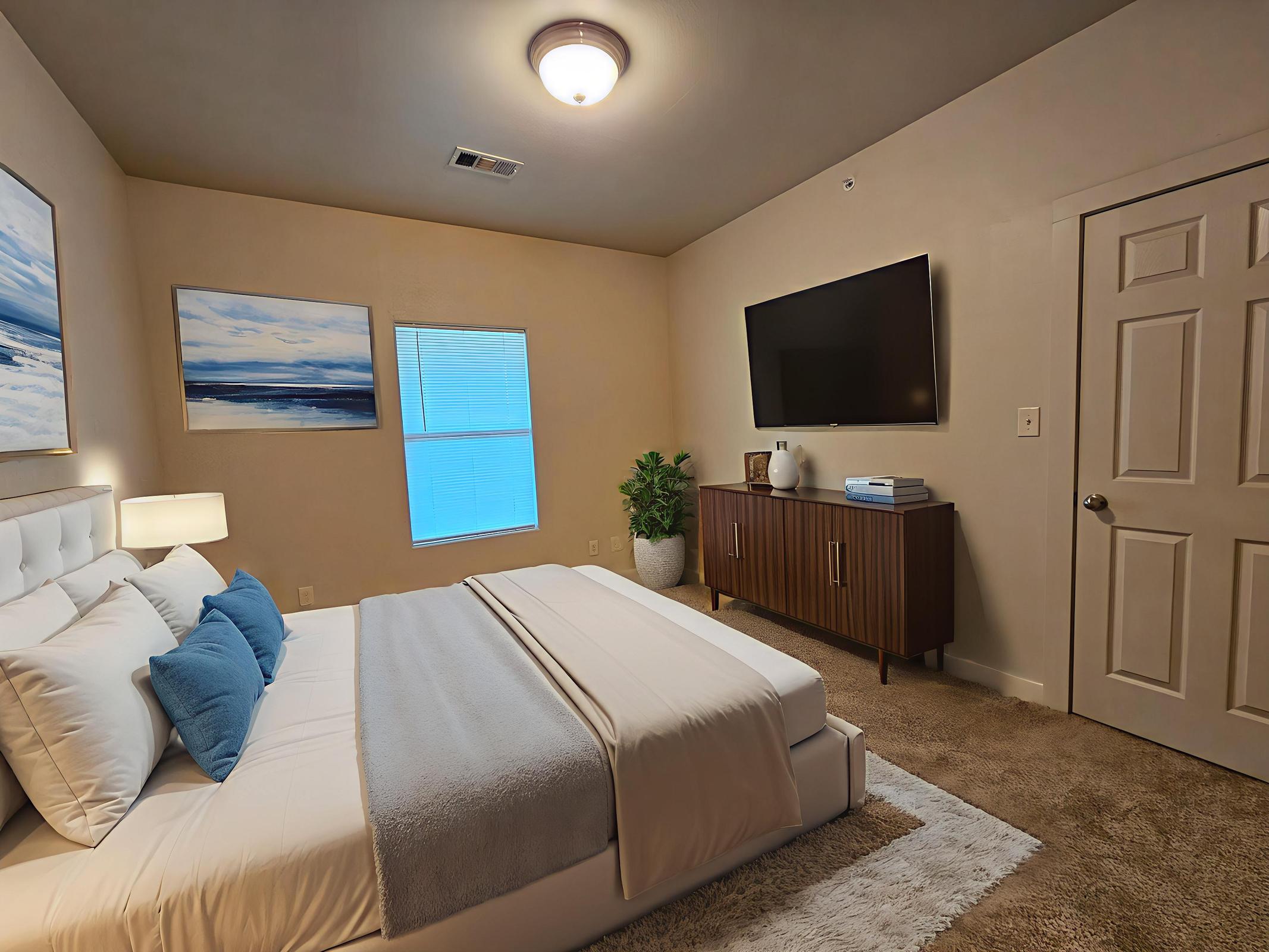 A cozy bedroom featuring a queen-sized bed with white linens and blue accent pillows. There are two abstract paintings on the walls, a window with light filtering through, a television mounted on the wall opposite the bed, and a wooden dresser beside a potted plant, all situated on plush carpet.