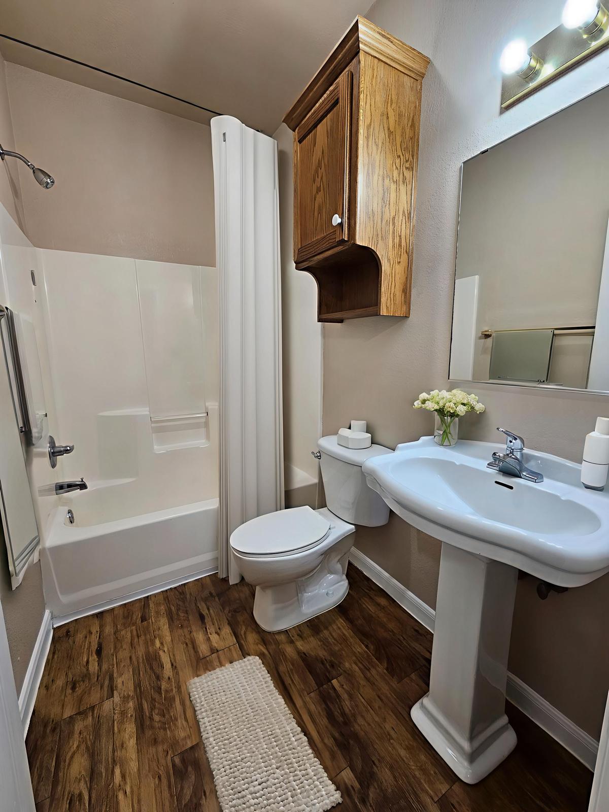 A clean and modern bathroom featuring a bathtub with a shower, a toilet, a pedestal sink with a mirror above, and a wooden cabinet. The flooring is a warm wood tone, and there are decorative elements like a small rug and a vase of flowers on the sink. The walls are painted in soft beige.