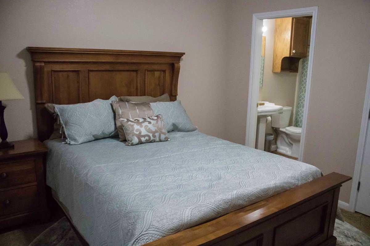 A cozy bedroom featuring a wooden bed frame with a light blue comforter and decorative pillows. A bedside table with a lamp is next to the bed. In the background, a doorway leads to a bathroom with a toilet and sink visible. The walls are a soft beige color, creating a warm atmosphere.