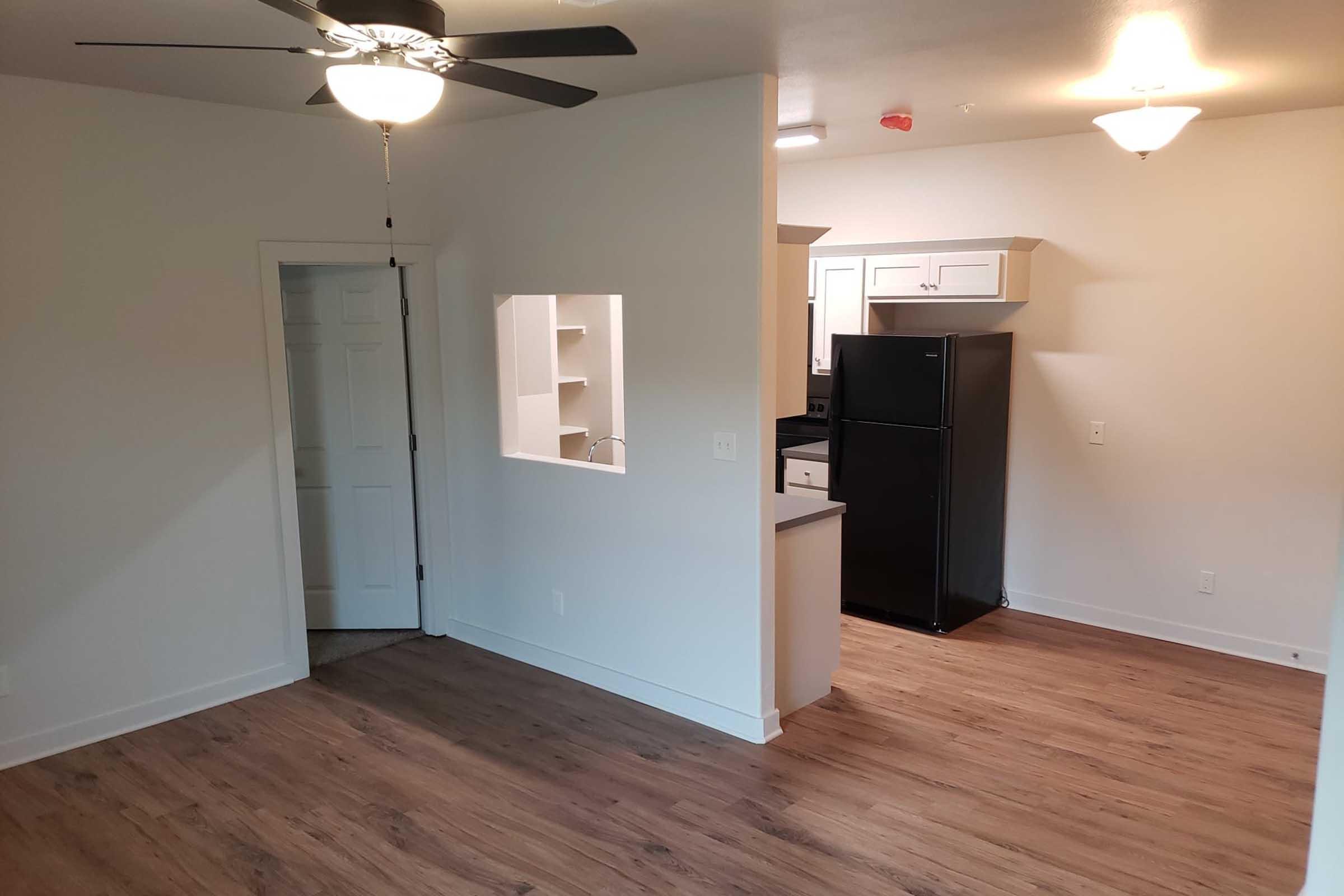A well-lit, modern apartment interior featuring a living area with wood-like flooring, a ceiling fan, and an open doorway leading to a kitchen area. The kitchen has white cabinetry and a black refrigerator visible. The walls are painted light, creating a spacious ambiance.