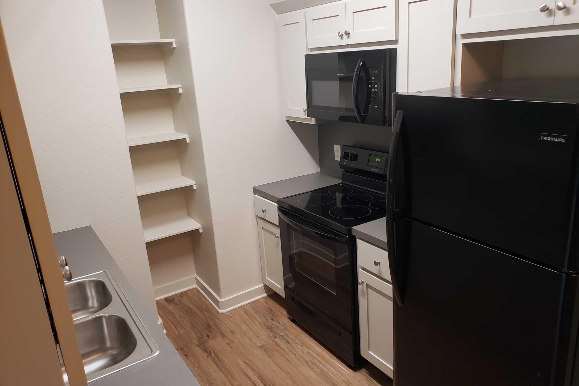 A small kitchen featuring white cabinets, a black stove, an over-the-range microwave, and a black refrigerator. There are two sinks on the counter, and a shelf for storage is visible on the wall. The flooring is a light wood laminate, creating a modern and tidy appearance.