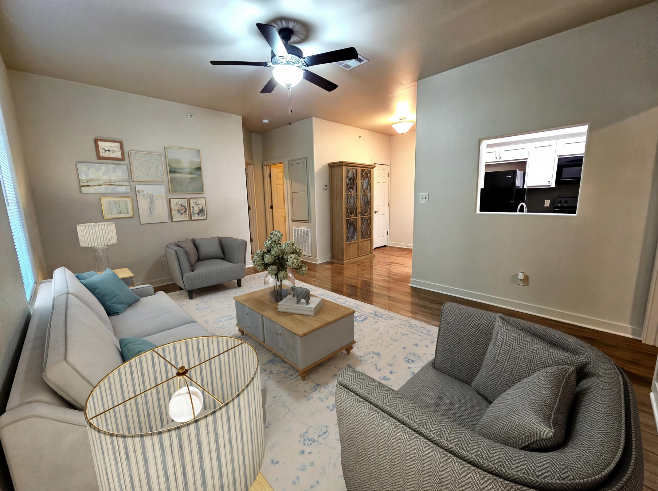 A modern living room featuring a light gray sofa and two armchairs, all arranged on a decorative rug. A coffee table sits center with a vase of flowers. Walls adorned with framed pictures, and a ceiling fan overhead. A hallway leads to a kitchen visible through an open wall. Soft natural light brightens the space.