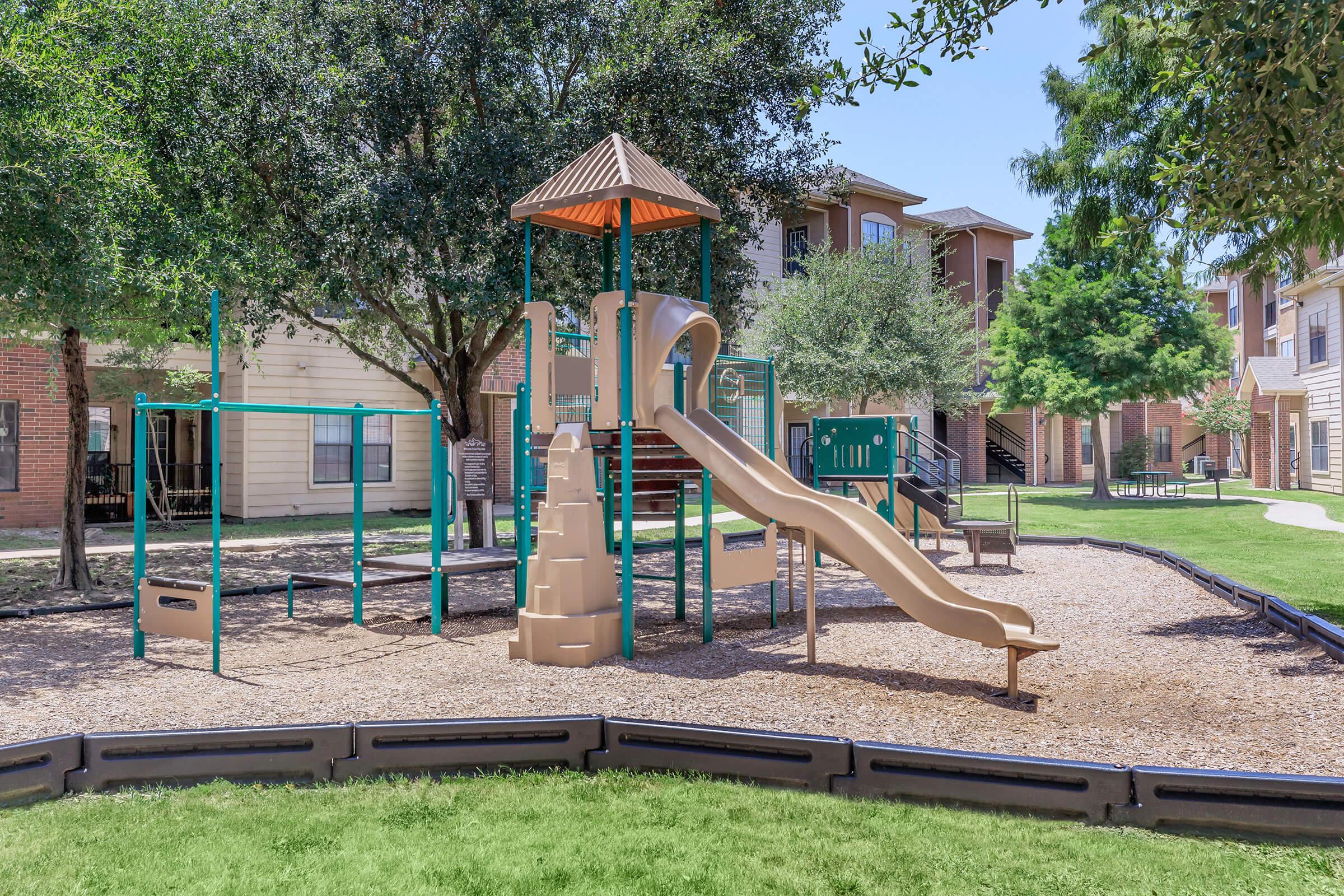 A playground featuring a tan slide and climbing structure surrounded by green grass and trees. The area includes a swing set and is located in a residential community with apartment buildings in the background. The setting is bright and inviting, suitable for children to play.
