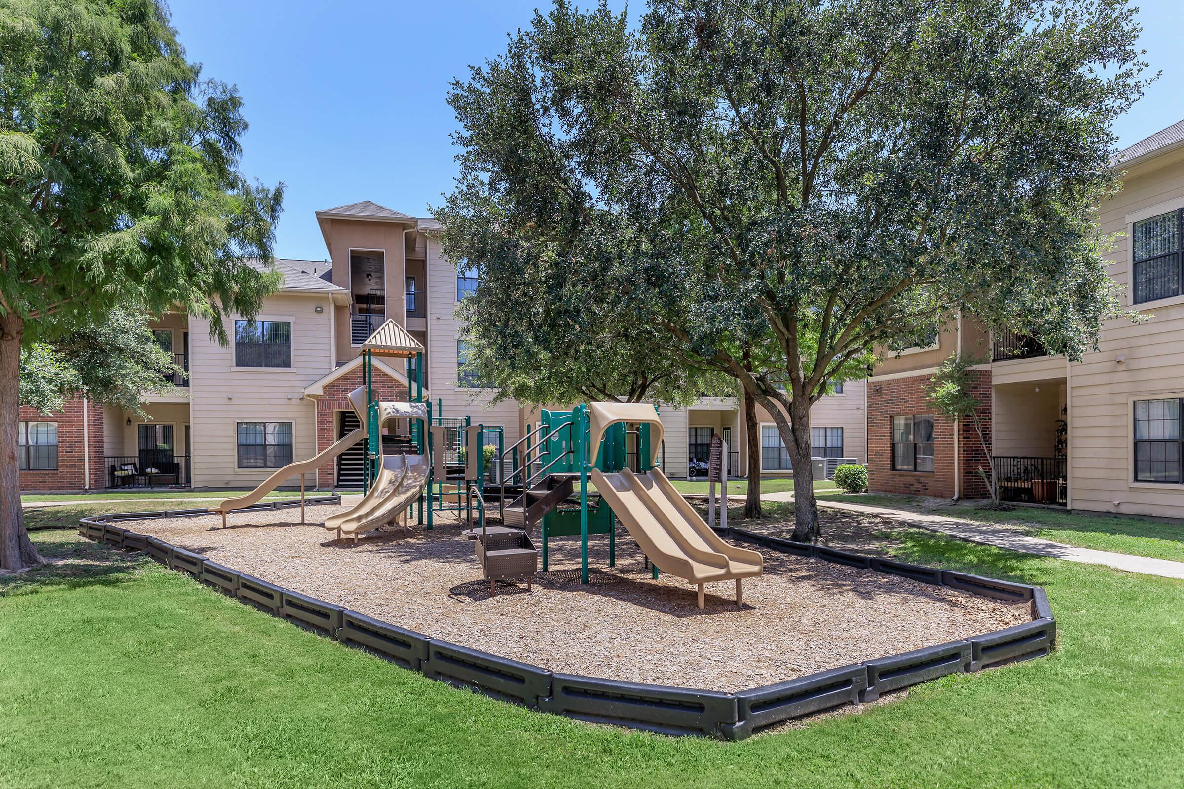 A playground with two slides and climbing structures surrounded by grassy areas and trees, located in a residential complex. Several apartment buildings are visible in the background, under a clear blue sky.