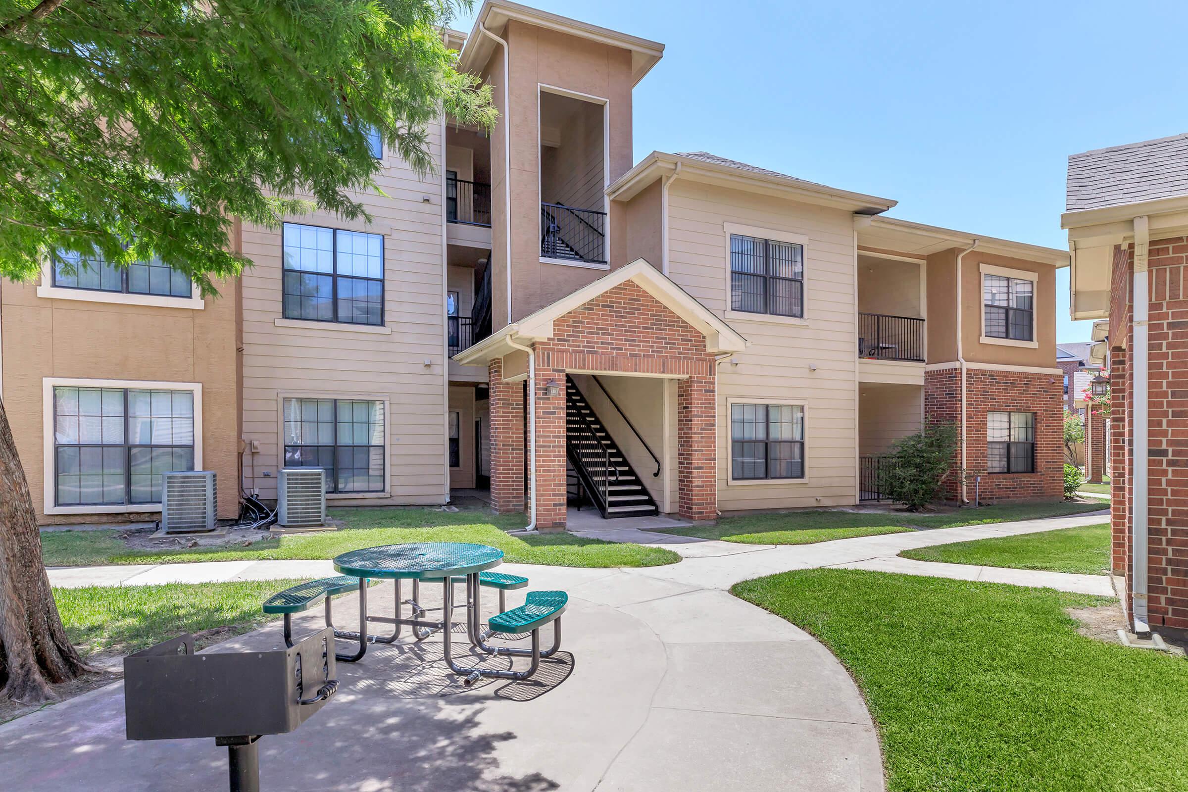 A view of an apartment complex featuring a two-story building with a brick entrance, surrounded by grass and trees. There are outdoor picnic tables and a barbecue grill in a shared courtyard area, with stairs leading up to the second floor of the building.