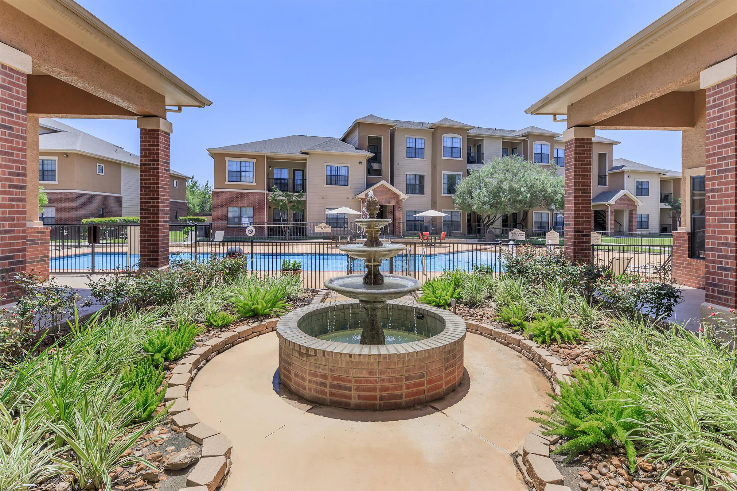 A peaceful courtyard featuring a circular brick fountain surrounded by plants, with a swimming pool and apartment buildings in the background under a clear blue sky.