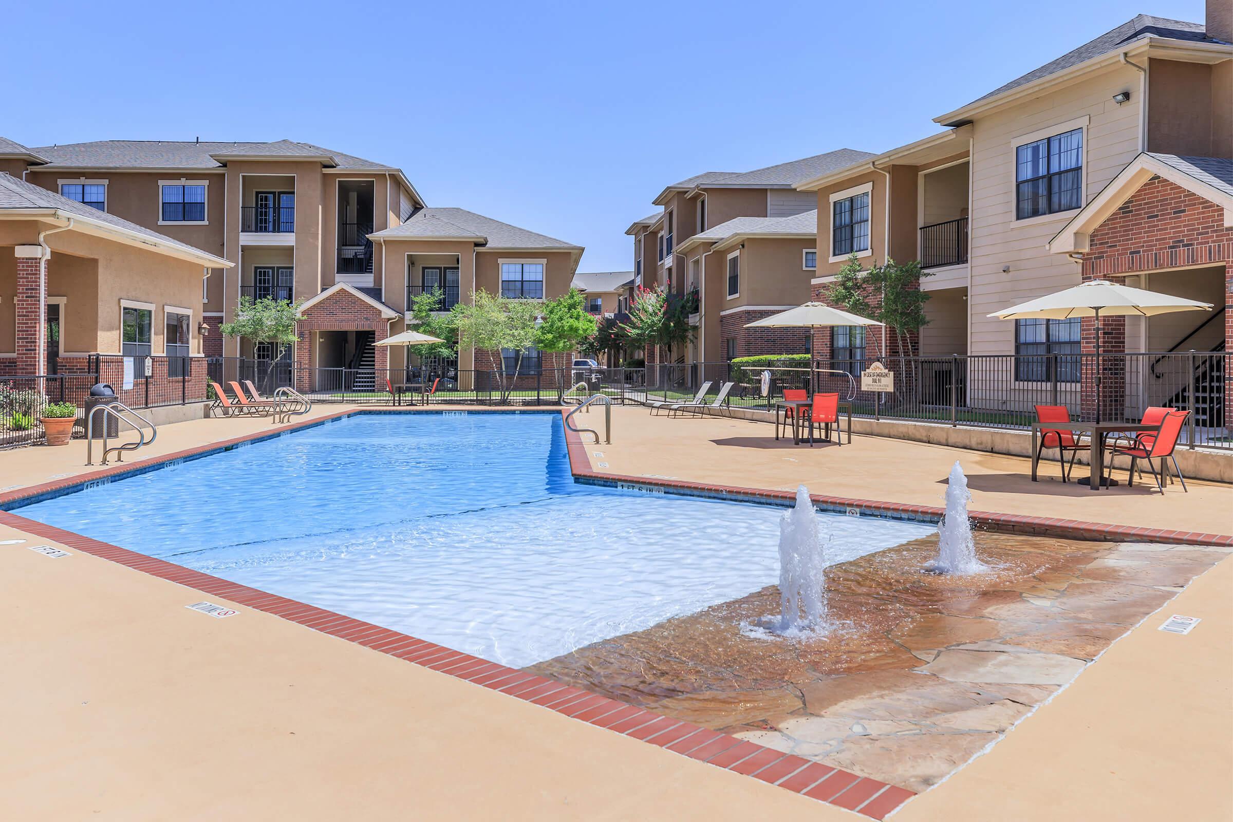 A clear blue swimming pool with fountains surrounded by a sunbathing area featuring lounge chairs. In the background, several apartment buildings with balconies and greenery can be seen. Red patio chairs and tables are placed near the pool, creating a relaxing outdoor atmosphere.