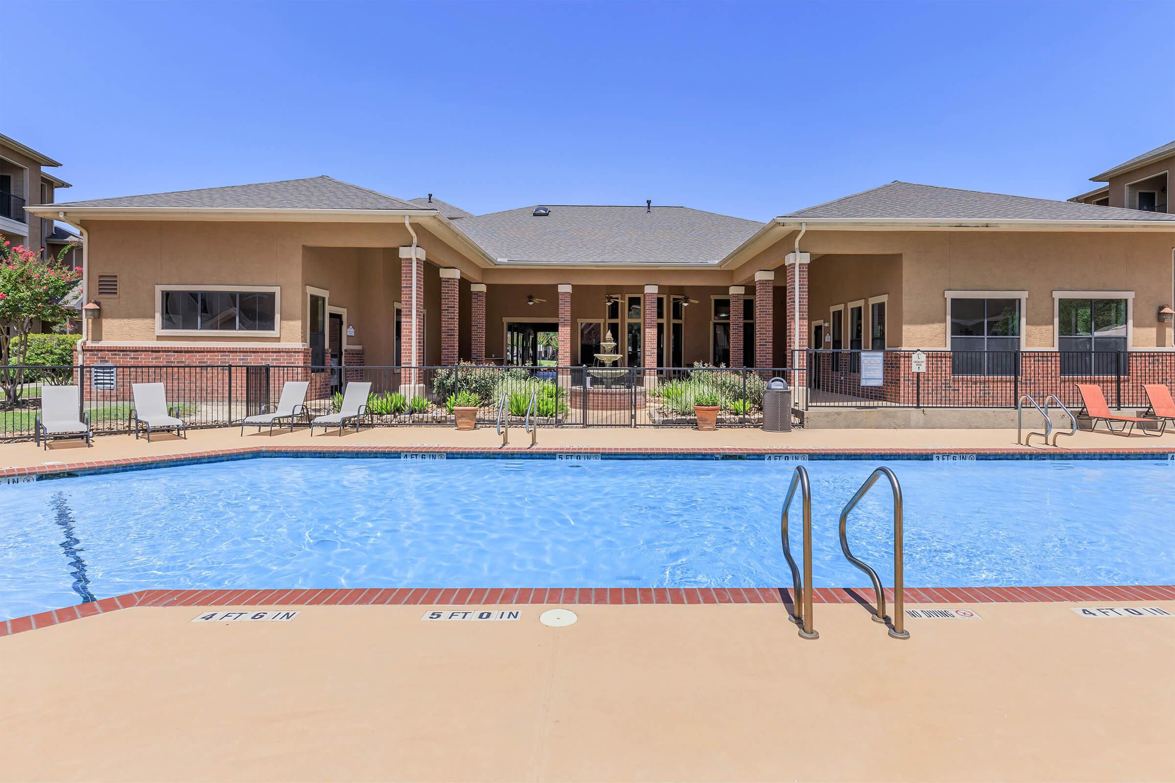A sparkling blue swimming pool with a ladder in the foreground, surrounded by lounge chairs. In the background, a shaded building with a covered porch and landscaped greenery, under a clear blue sky. The area appears inviting and well-maintained, ideal for relaxation and leisure.