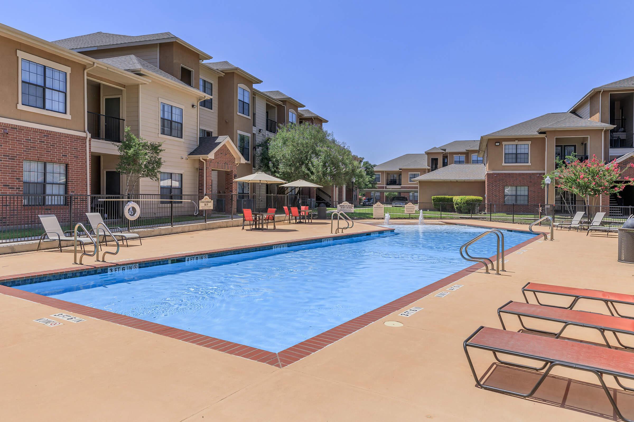 A clean, inviting swimming pool surrounded by a landscaped courtyard, with sun loungers and patio tables under umbrellas. Nearby, two multi-story apartment buildings are visible against a clear blue sky, creating a relaxed residential atmosphere.