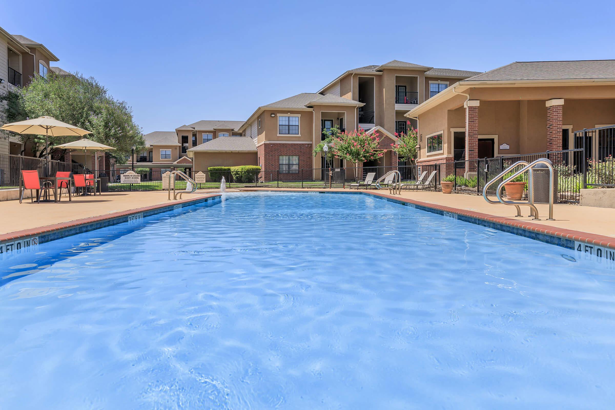 A clear blue swimming pool surrounded by lounge chairs and umbrellas, with apartment buildings visible in the background under a bright blue sky. The pool area is well-maintained, featuring a shallow end marked with a depth indicator. Lush greenery adds a refreshing touch to the scene.