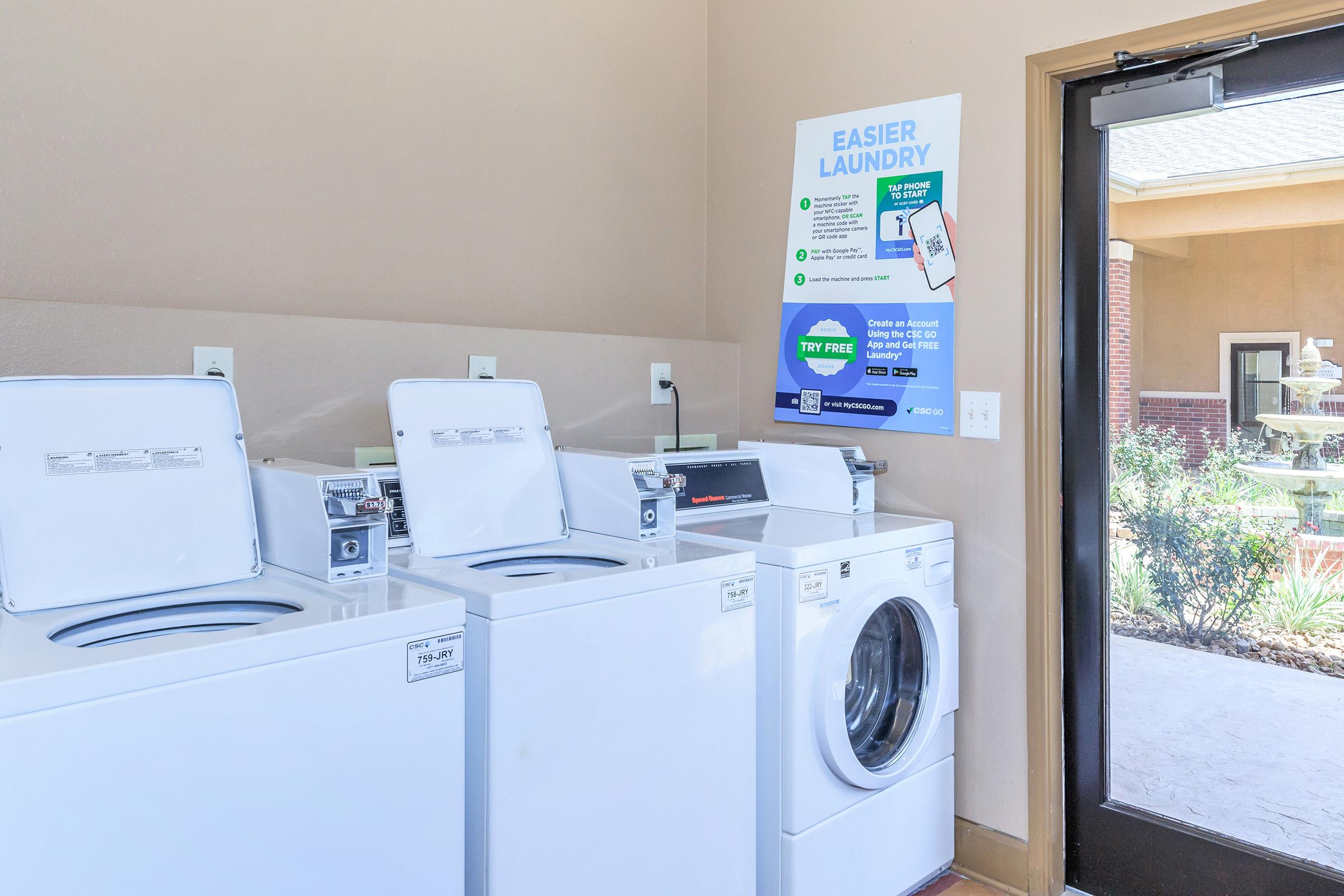 A laundry room featuring two white washing machines and a white dryer. A sign on the wall highlights the benefits of using the laundry service and promotes a free trial. A glass door leads to an outdoor area with plants and natural light.
