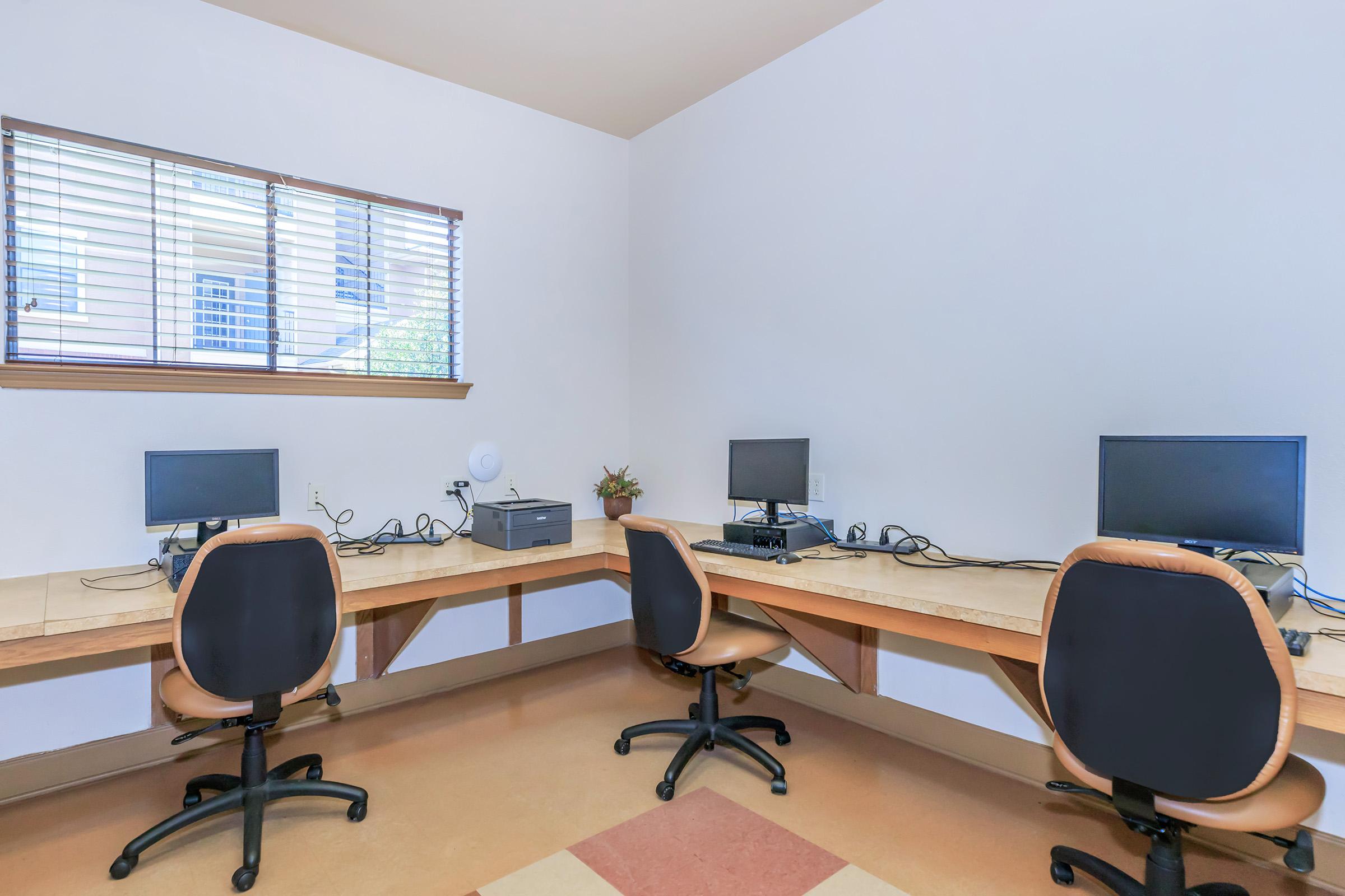 A workspace featuring two wooden desks with computers and black office chairs. Each desk has a monitor, keyboard, and mouse. A small printer is also on one desk, and there’s a window with blinds letting in natural light. The room has a clean, minimalistic design with light-colored walls.