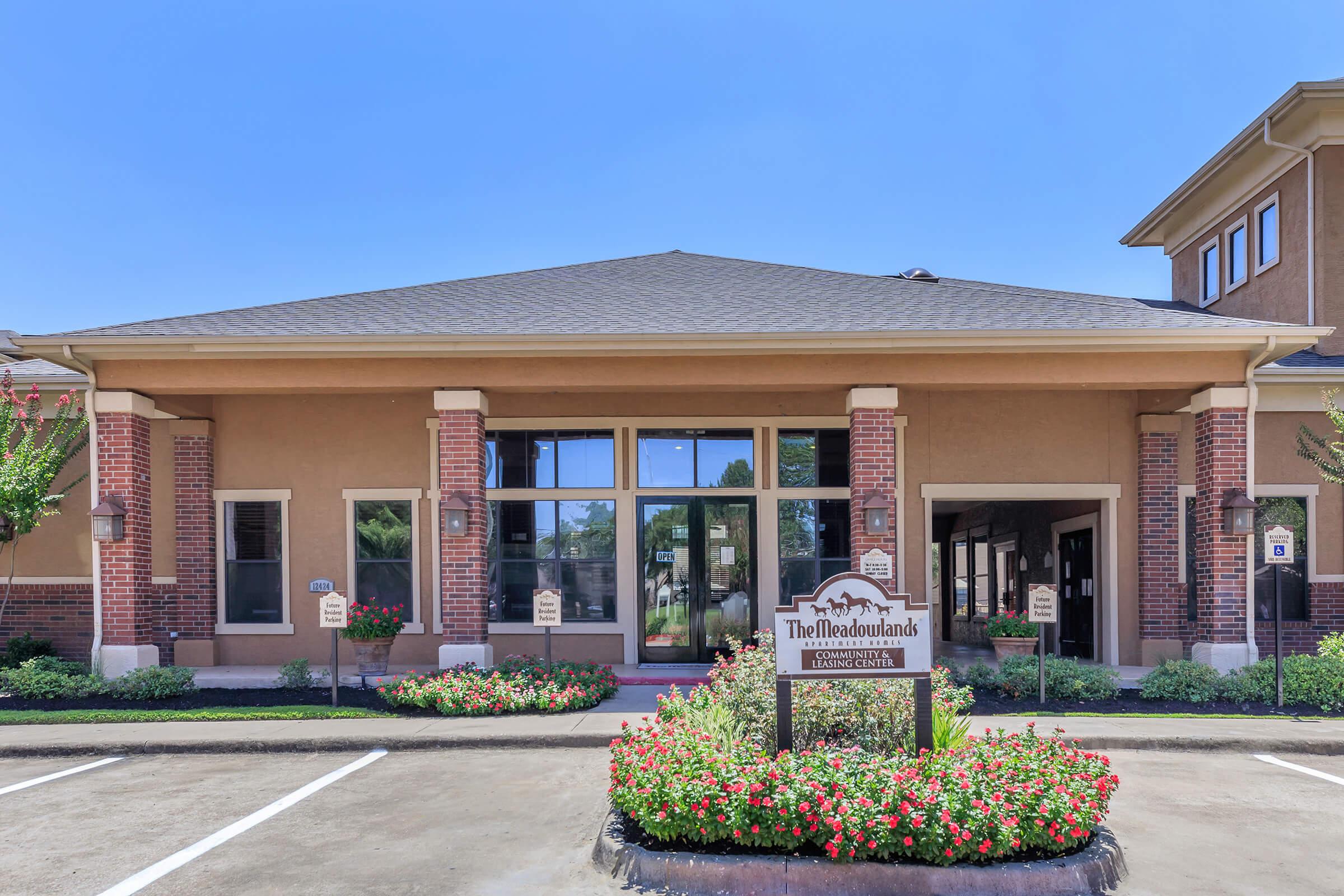 Front view of a building with a sign reading "The Woodlands" prominently displayed. The entrance is flanked by flowerbeds and well-maintained landscaping. Clear blue sky overhead, with a parking area visible in front. The architectural style features brick and subtle decorative elements.