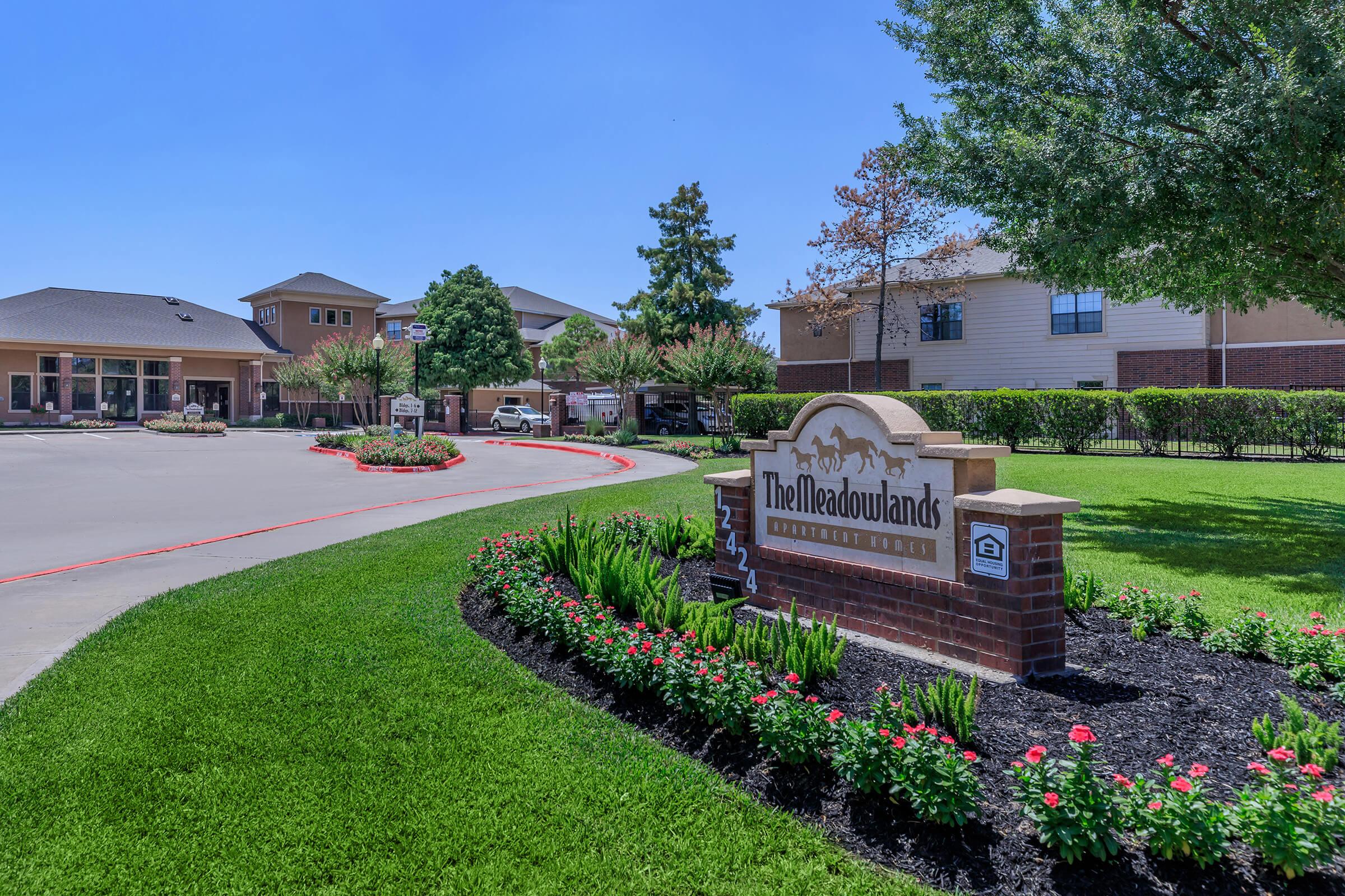 A welcoming entrance sign for The Meadowlands apartment community, surrounded by a manicured lawn with vibrant flowers and shrubs. In the background, various residential buildings are visible under a clear blue sky.