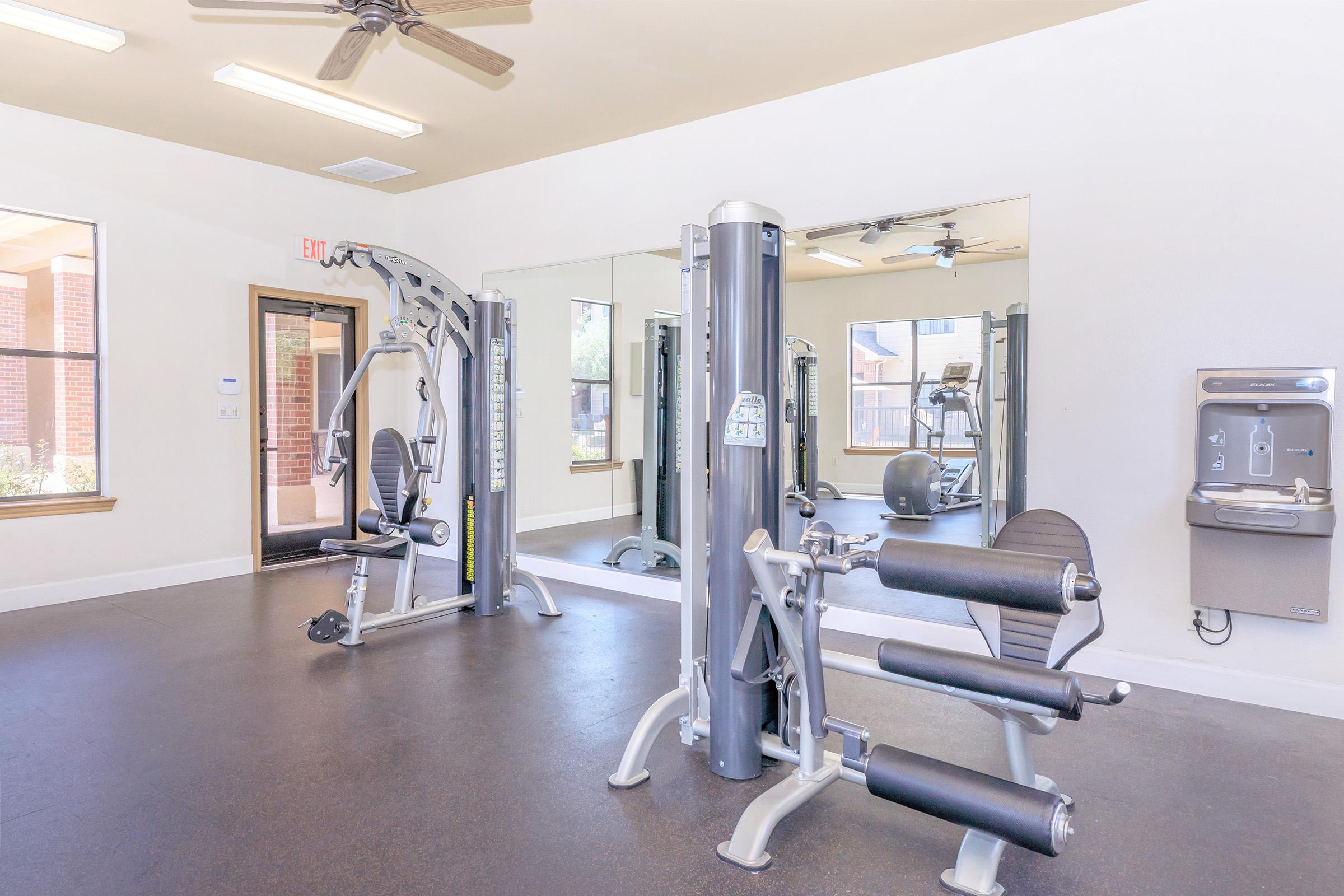 A bright and spacious gym area featuring various exercise equipment, including a multi-gym machine and exercise bike. Large windows provide natural light, and mirrors line one wall. A water fountain is visible in the background, enhancing the workout space's functionality.