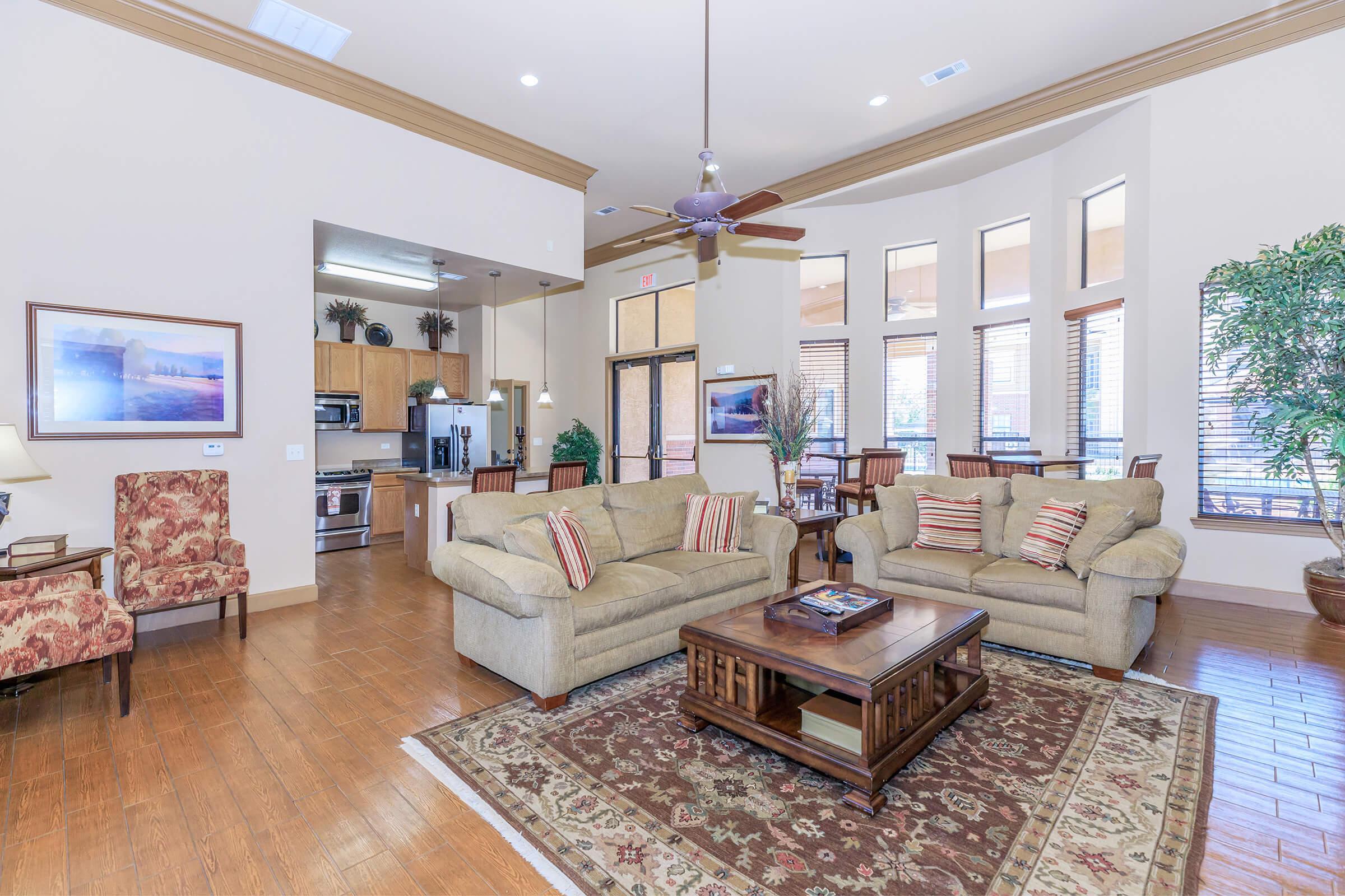 A cozy living area featuring a beige sofa and armchairs with decorative cushions, a wooden coffee table, and a carpet. Large windows allow natural light, and a dining area is visible in the background. The decor includes plants and artwork on the walls, creating a warm and inviting atmosphere.