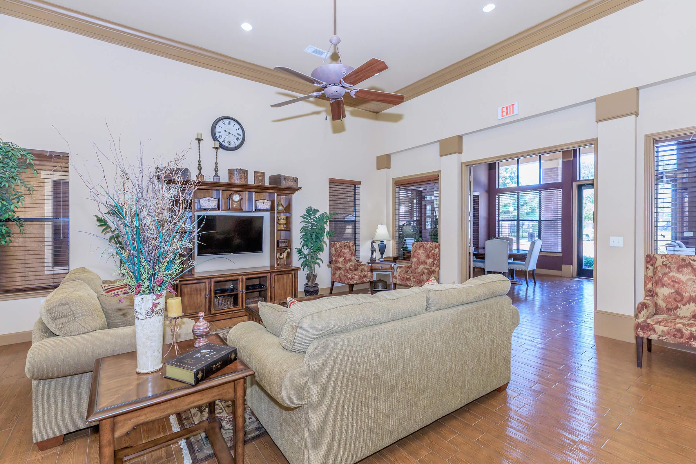 A cozy living room with a beige sofa and armchairs, a wooden coffee table, and a television unit. The room features warm lighting, a ceiling fan, decorative plants, and large windows with wooden blinds, creating a welcoming atmosphere. Wall art and a clock add finishing touches to the space.