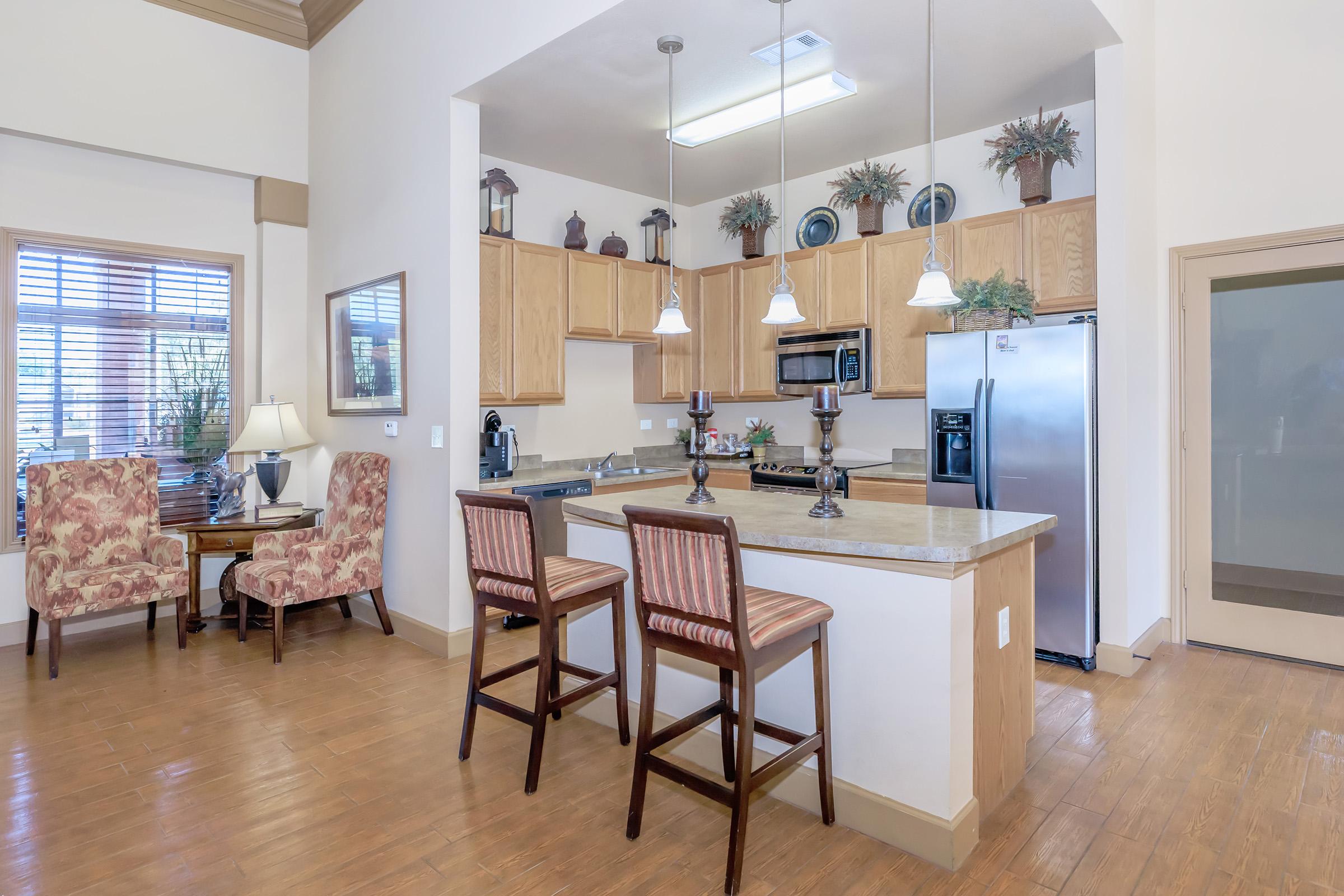 A modern kitchen featuring wooden cabinetry, stainless steel appliances, and a spacious island with bar stools. The dining area includes a small table and chairs, while the room is brightened by natural light from large windows and decorative plants. The flooring is a warm hardwood.