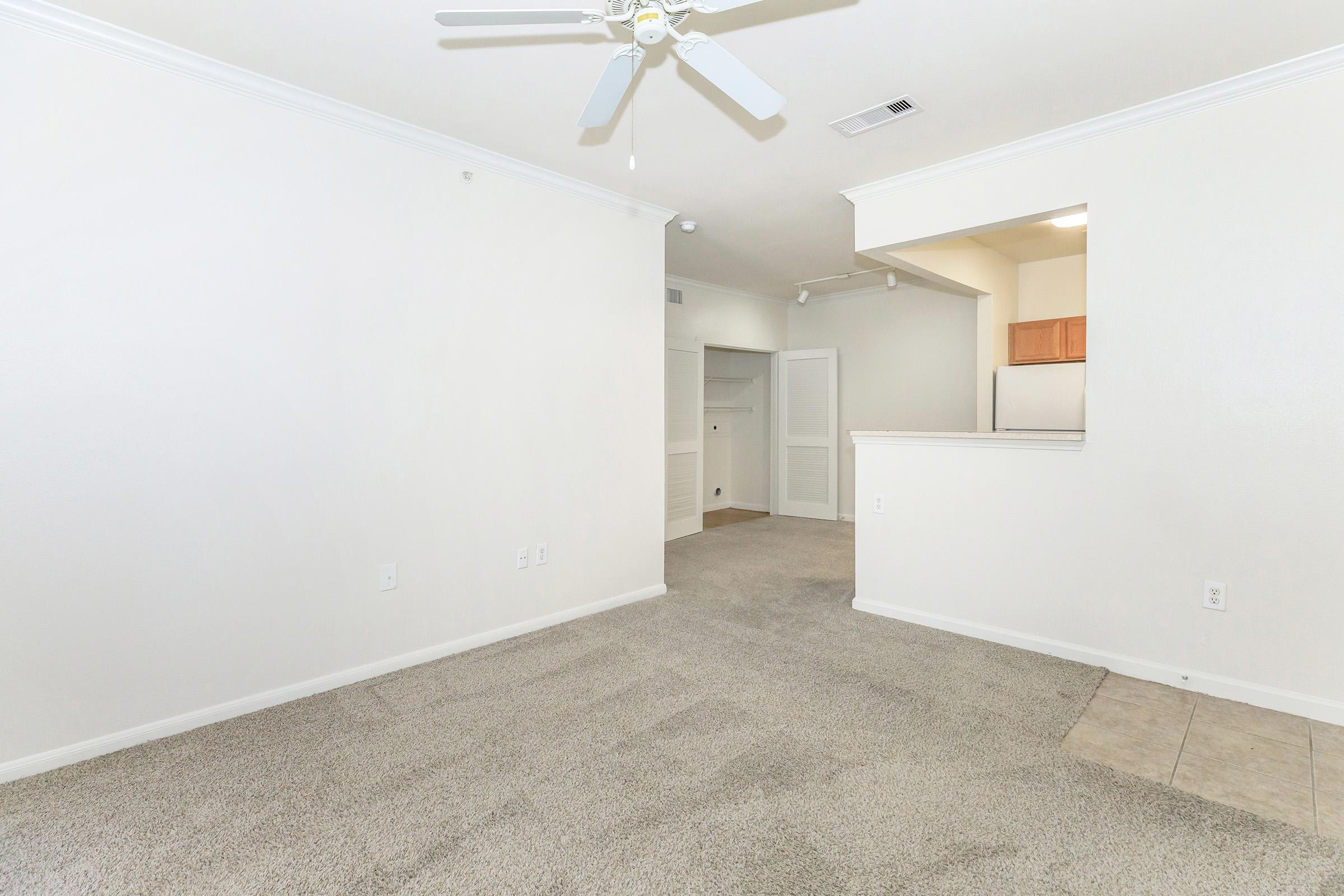 A spacious, light-colored living area with carpet flooring, featuring a ceiling fan and an open layout. The view includes an archway leading to a kitchen area with wooden cabinets visible in the background, along with a door to a hallway, creating an airy and inviting atmosphere.