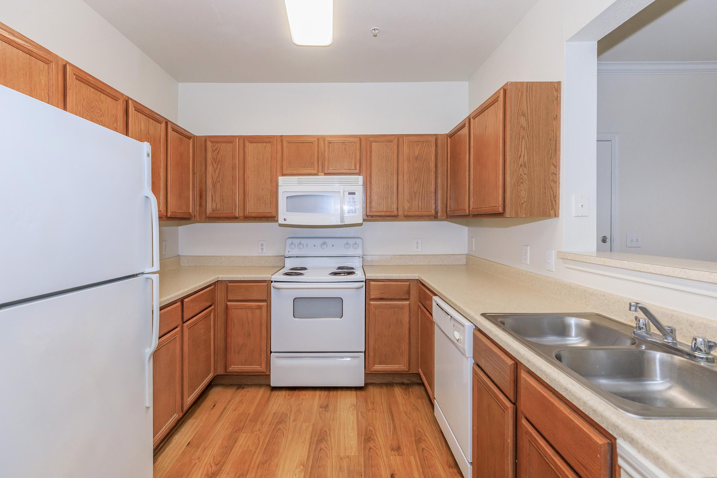 A modern kitchen featuring wooden cabinets, a white refrigerator, a microwave above a white stove, and a dishwasher. The countertop is light-colored, and there are double sinks. The flooring is wood, and the walls are painted in a neutral tone. The kitchen is well-lit with a ceiling light.