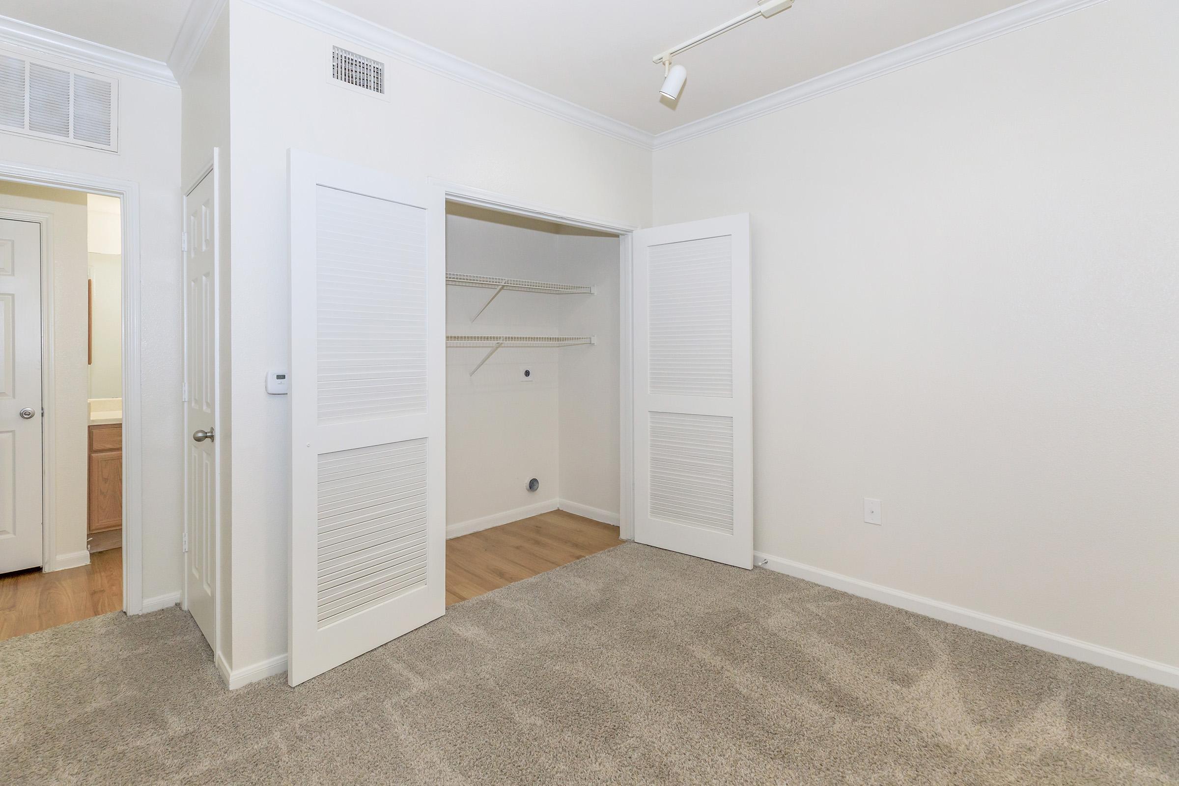 Empty room with light-colored walls and carpet. It features a double-door closet with shelves and a small opening for plumbing. There is a doorway leading to a bathroom visible in the background. The room is well-lit with a light fixture on the ceiling.