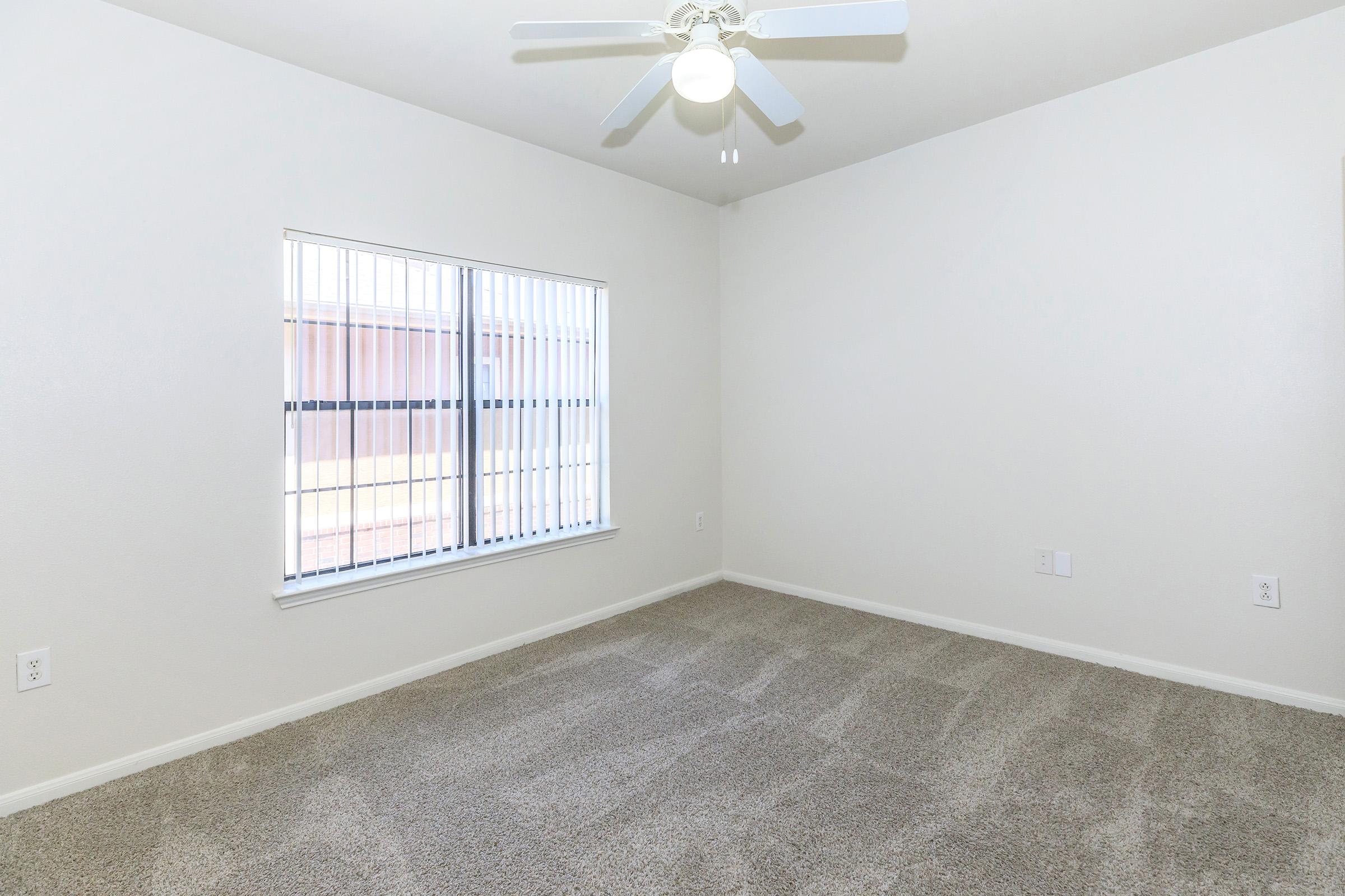 A vacant, well-lit room featuring light-colored walls and plush beige carpet. A ceiling fan is mounted in the center, and a large window with blinds allows natural light to fill the space. The room is empty, creating a spacious and inviting atmosphere.